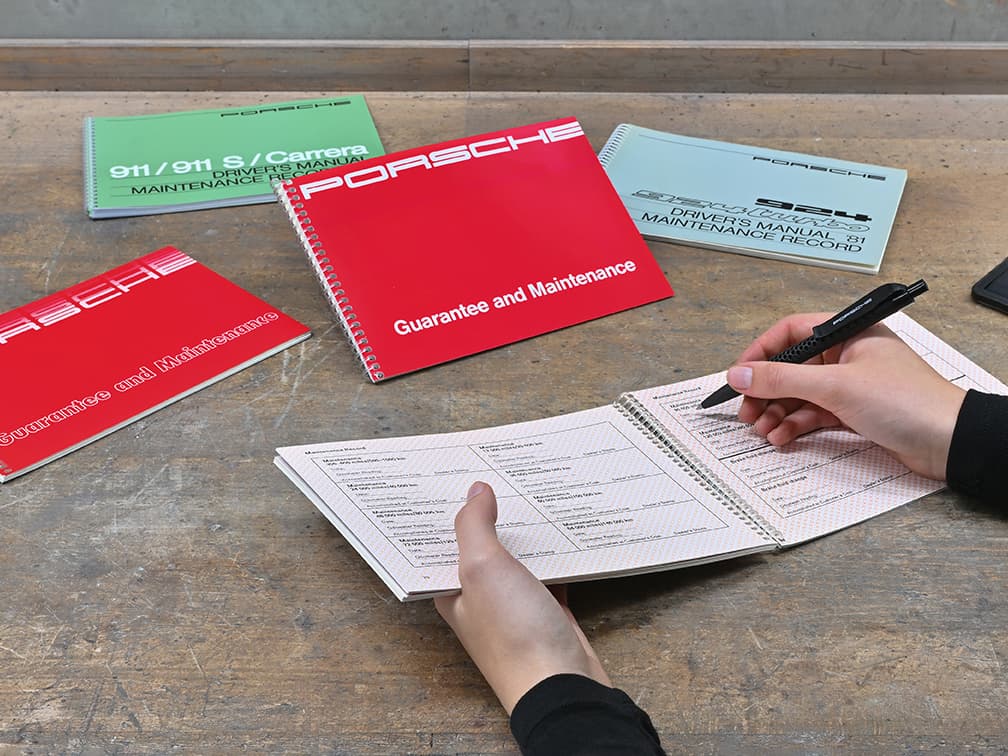 Hands writing in a maintenance record book on a wooden table, surrounded by Porsche manuals in red, green, and blue covers.
