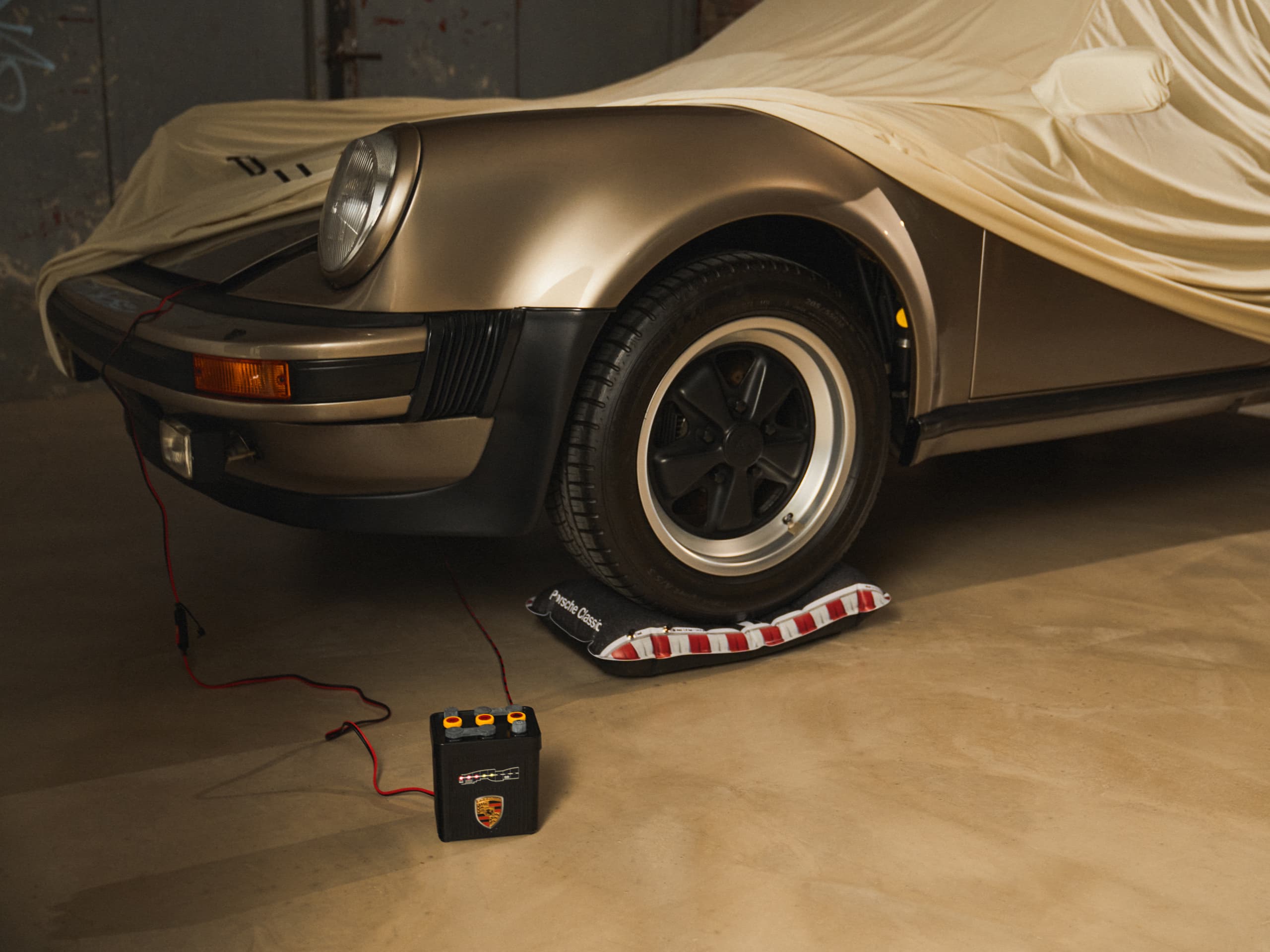 Classic Porsche partially covered, gold color, with a battery charger connected, parked on a concrete floor.