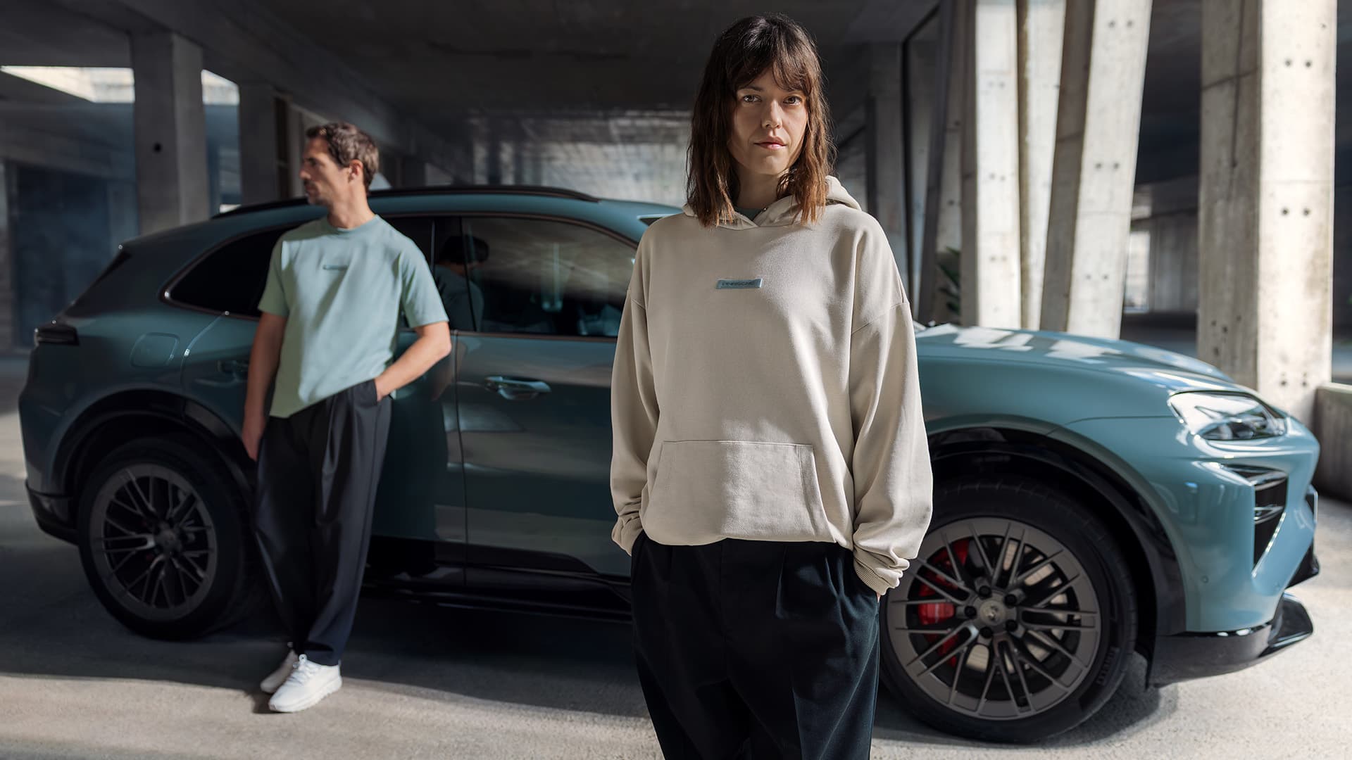 Woman in beige hoodie and man in blue shirt stand by a blue Porsche in a concrete garage, sunlight streaming in.