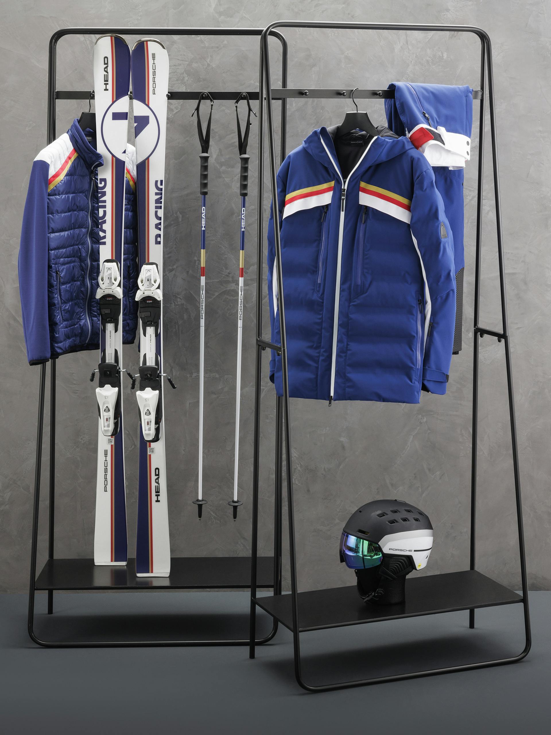 Various products from the new Porsche HEAD Ski collection 2023 pictured hanging on two small clothing racks against a plain grey background.