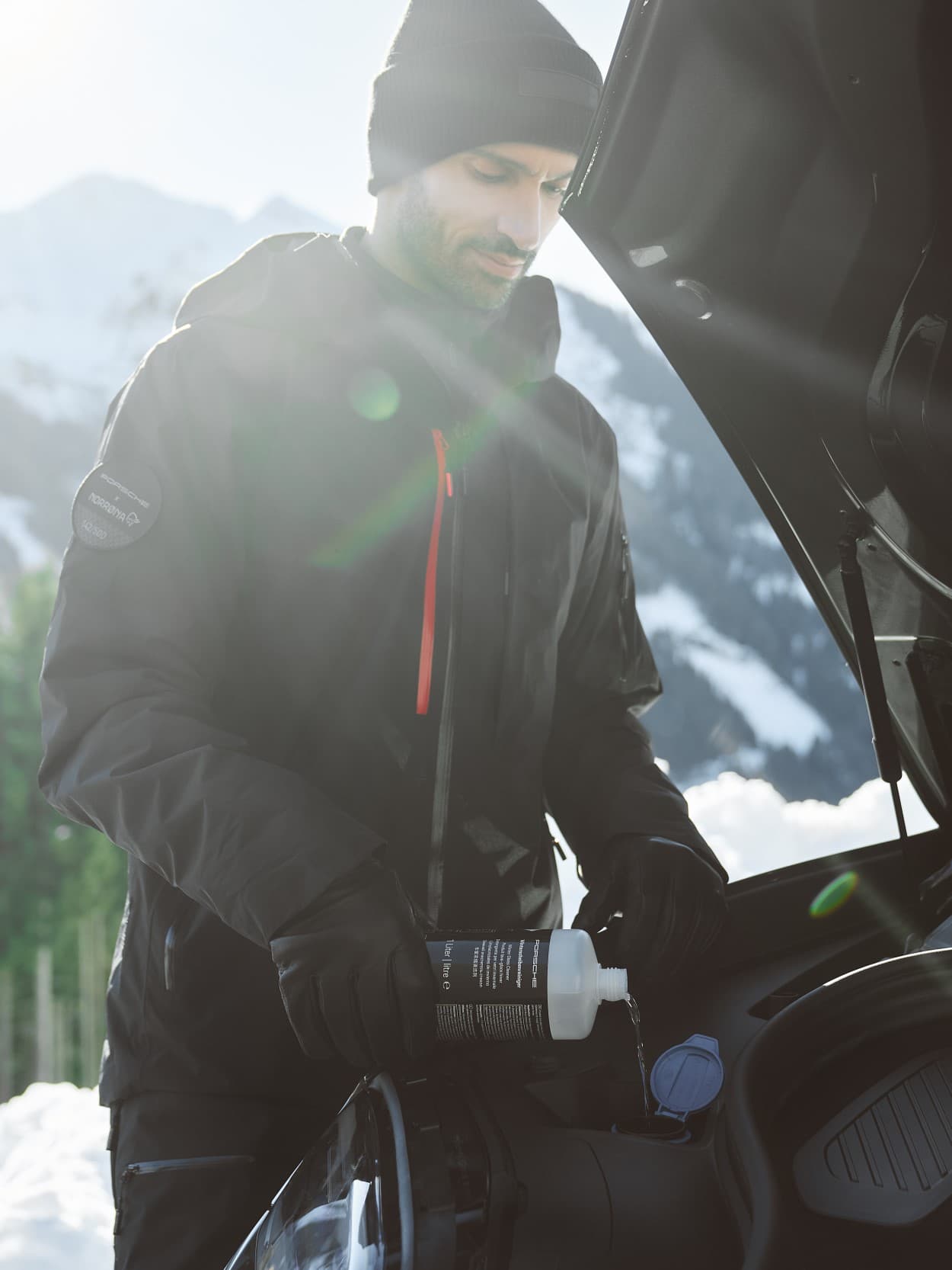 Man in black jacket and beanie pouring liquid into car engine, snowy mountains in background.