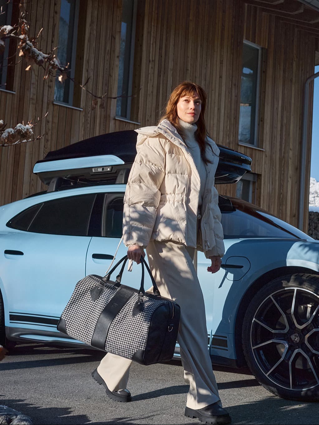 Woman in beige coat with checkered bag walks by a blue car, wooden building in background, winter setting