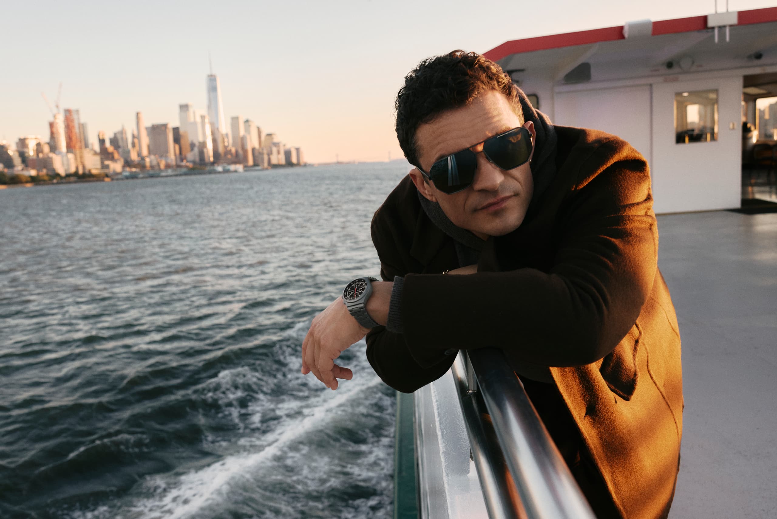 “Orlando Bloom wears the Porsche Design Targa Eyewear series and leans against the railing of a ferry, overlooking the water with the city skyline of New York in the distance.
