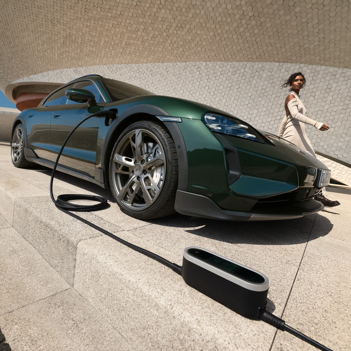 Green Porsche Taycan charging on a concrete platform, with a woman in a beige dress walking nearby.