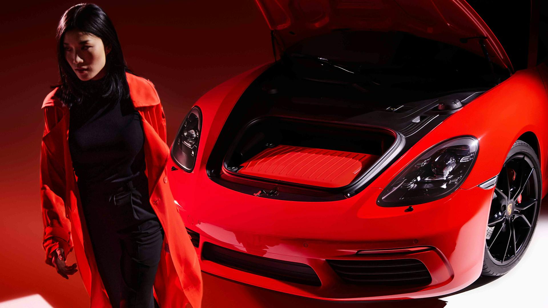 Woman wearing black clothing and a red coat walking away from a red Porsche 911, containing a red Porsche Design Hardcase Trolley in the front luggage compartment.