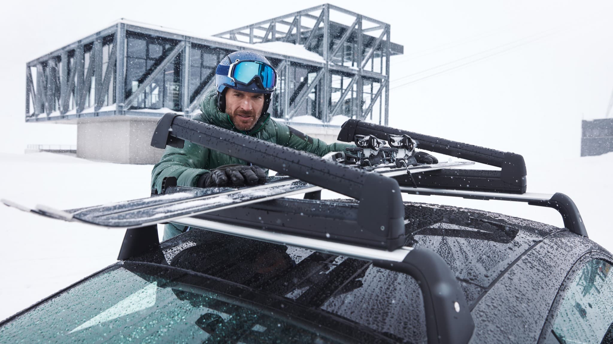 Mann in grüner Jacke und blauem Helm befestigt Skier auf dem Dachgepäckträger eines Autos in verschneiter Landschaft mit modernem Gebäude im Hintergrund.