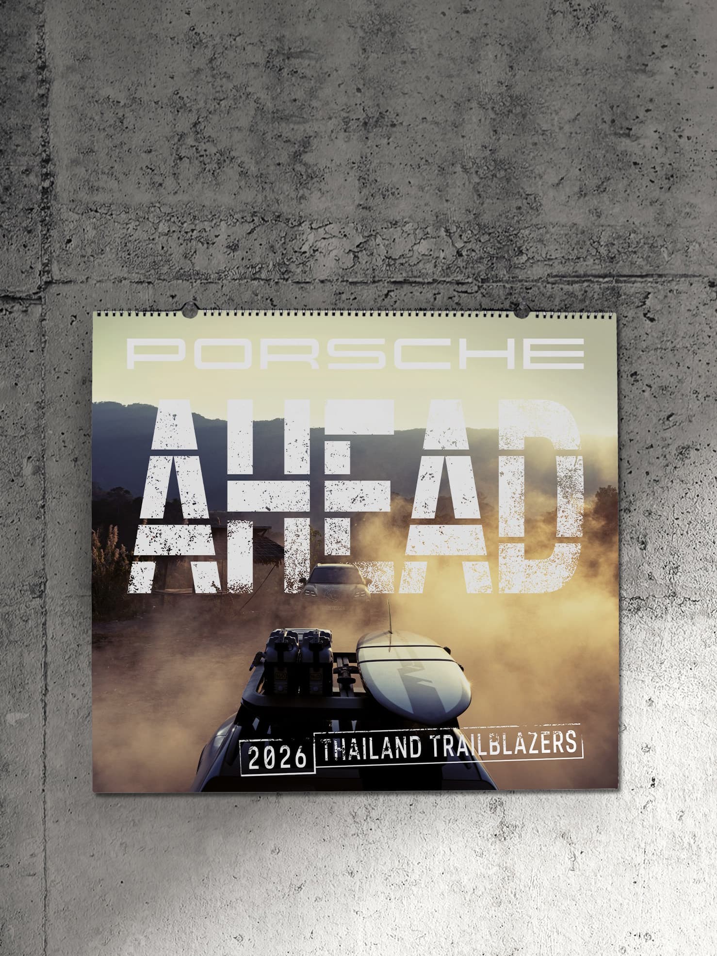 Porsche calendar on concrete wall, featuring "AHEAD" text, car with surfboard, and "2026 Thailand Trailblazers" theme.