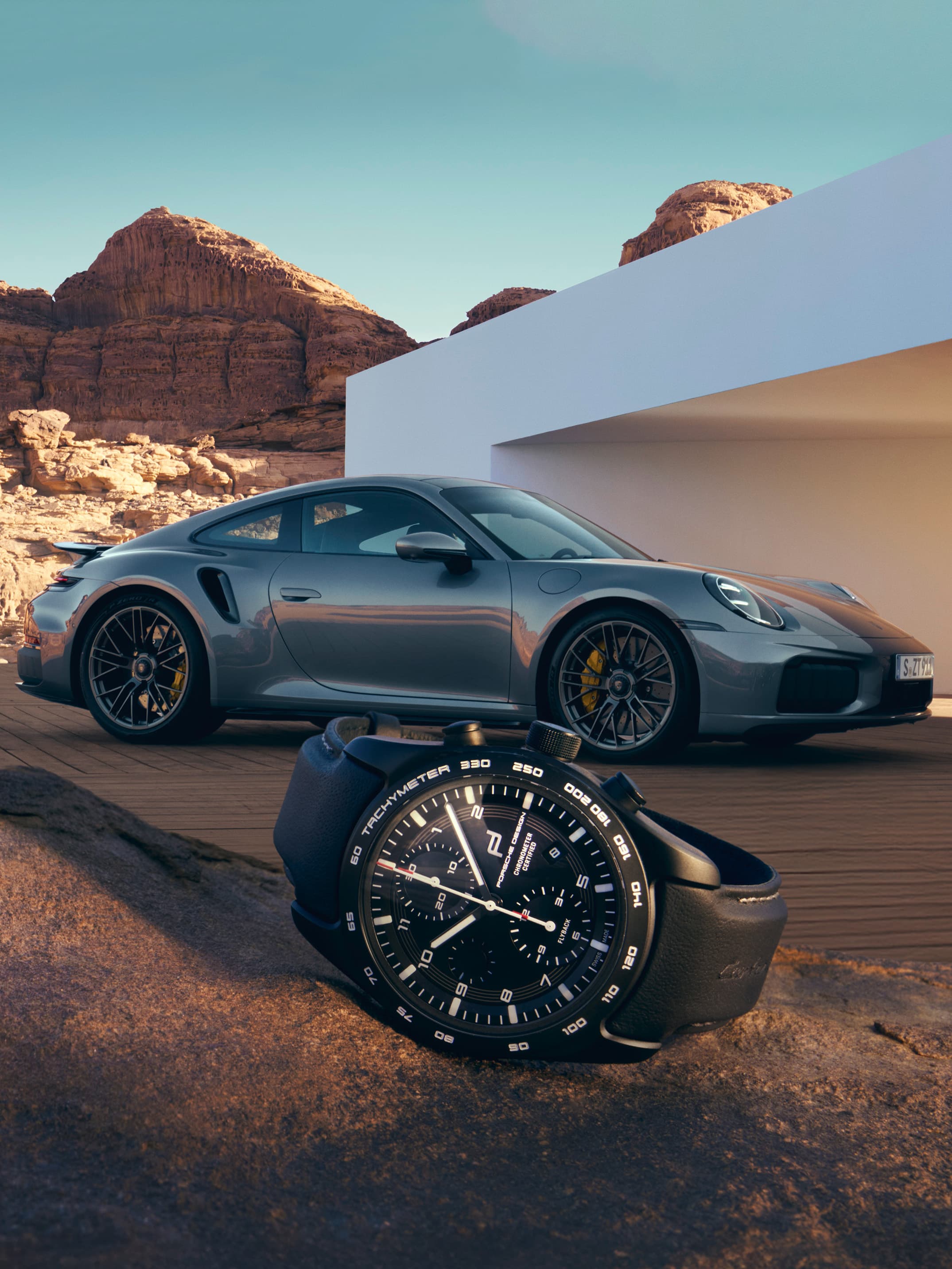 Schwarze, elegante Uhr auf felsigem Untergrund, mit silbernem Porsche-Sportwagen und Wüstenlandschaft im Hintergrund.