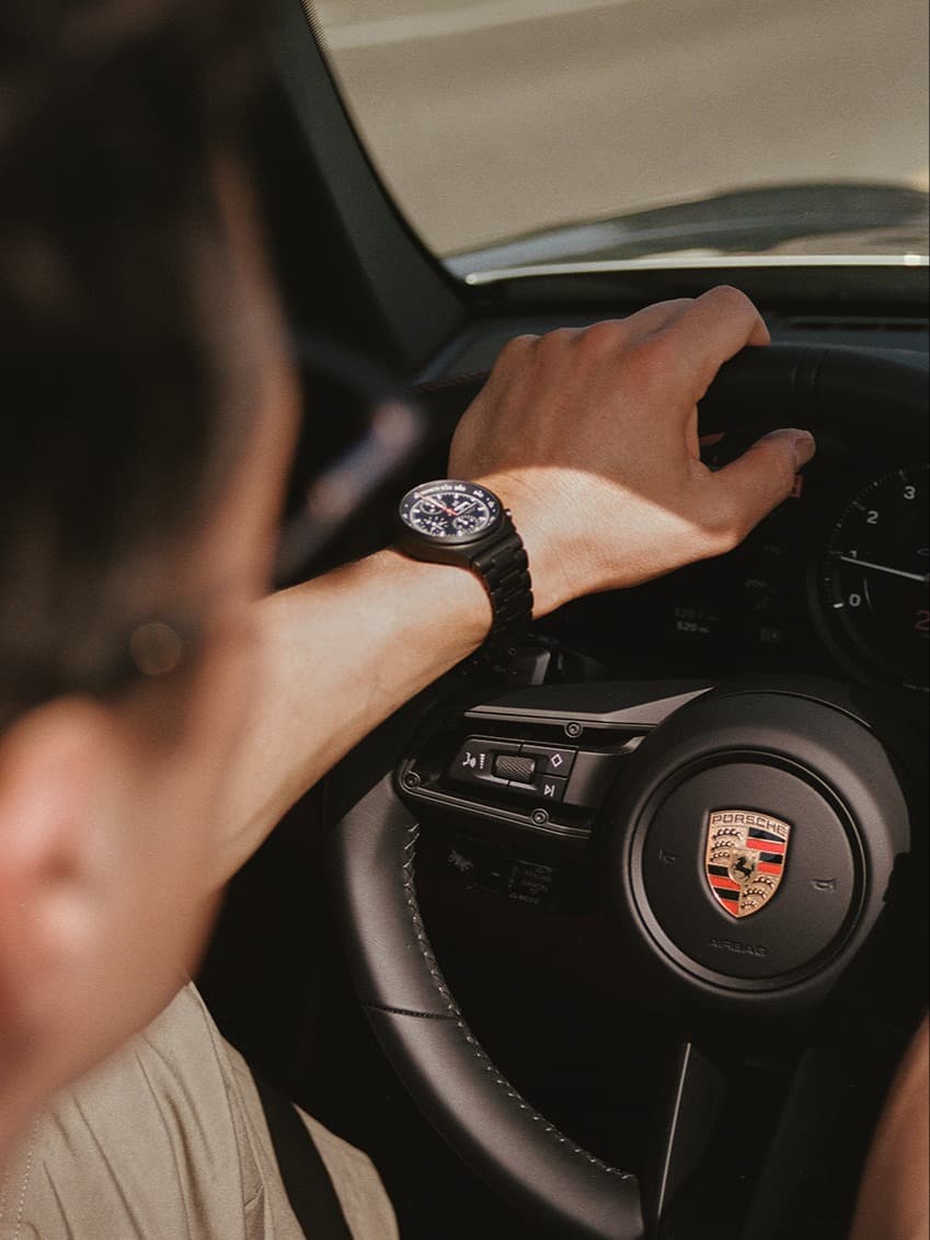 Hand op Porsche-stuurwiel, met een zwart horloge, gezien vanuit de auto.