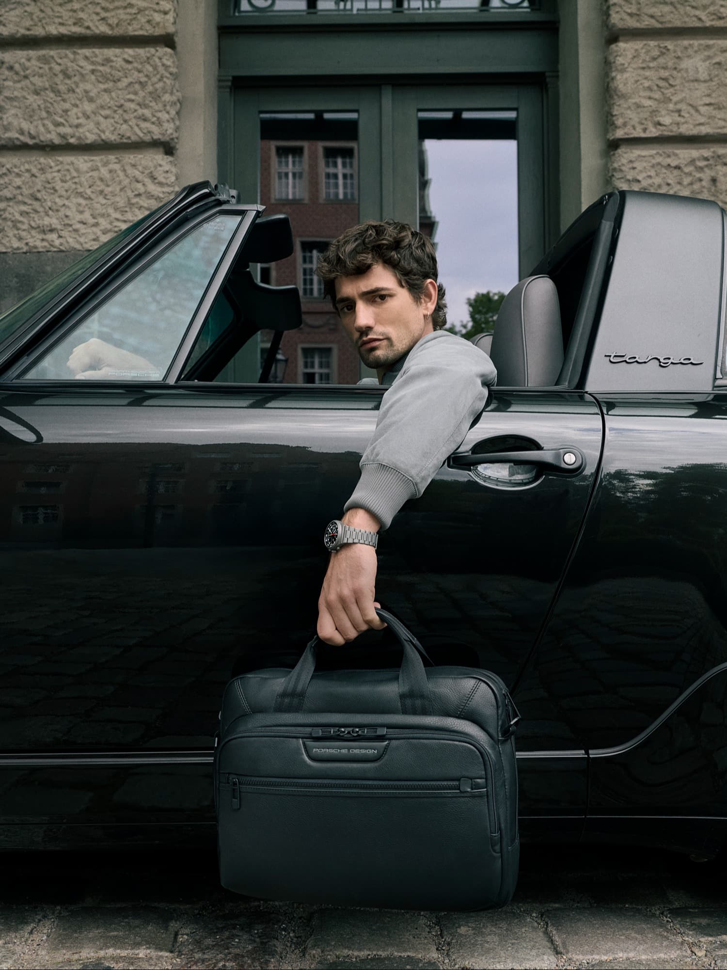 Man in a car holding a black Porsche Design bag, wearing a watch, with a city building in the background.