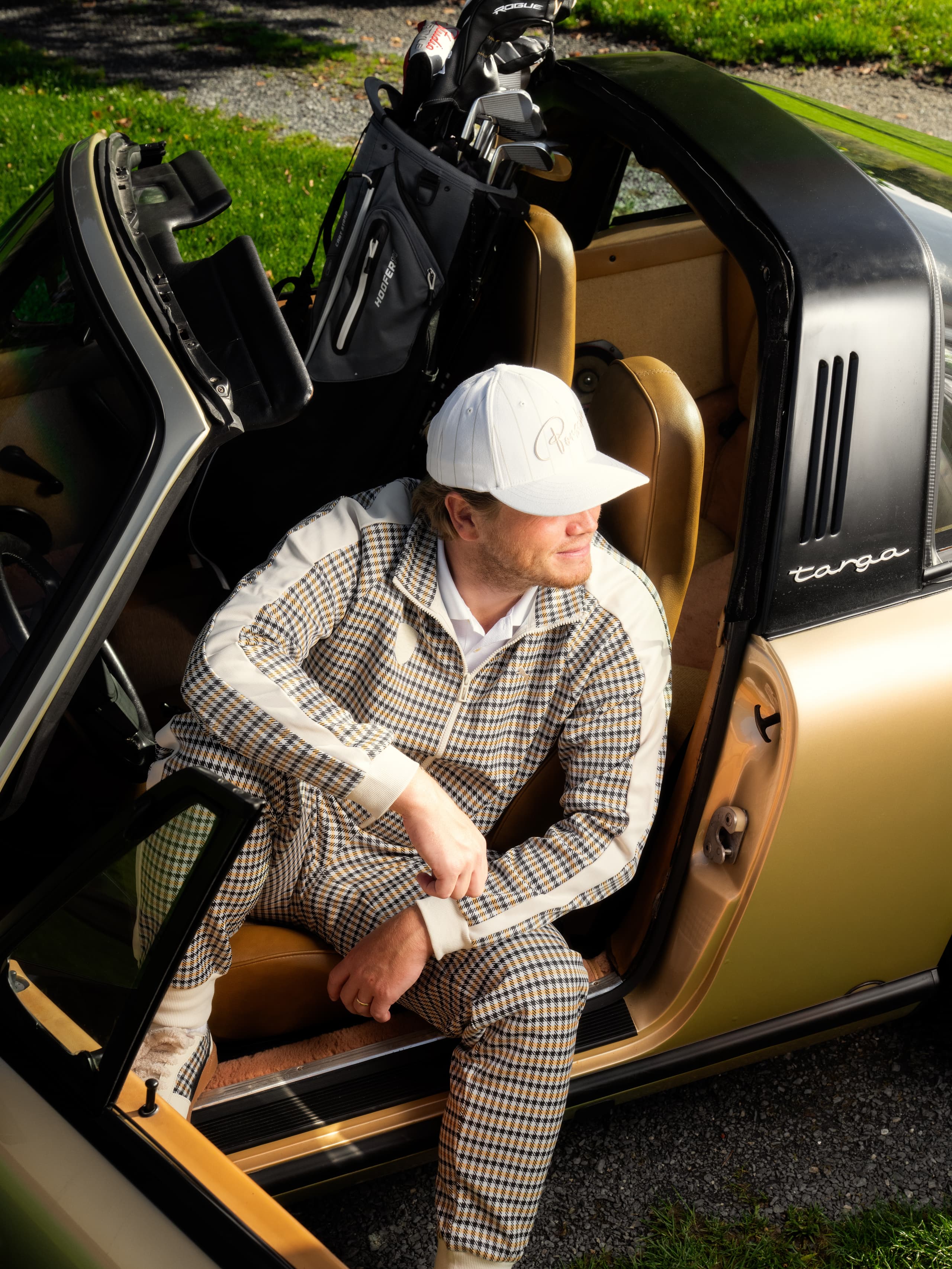 Mann in kariertem Outfit und weißer Mütze sitzt in einem goldenen Porsche Targa mit Golfschlägern im Heck, auf Gras geparkt.
