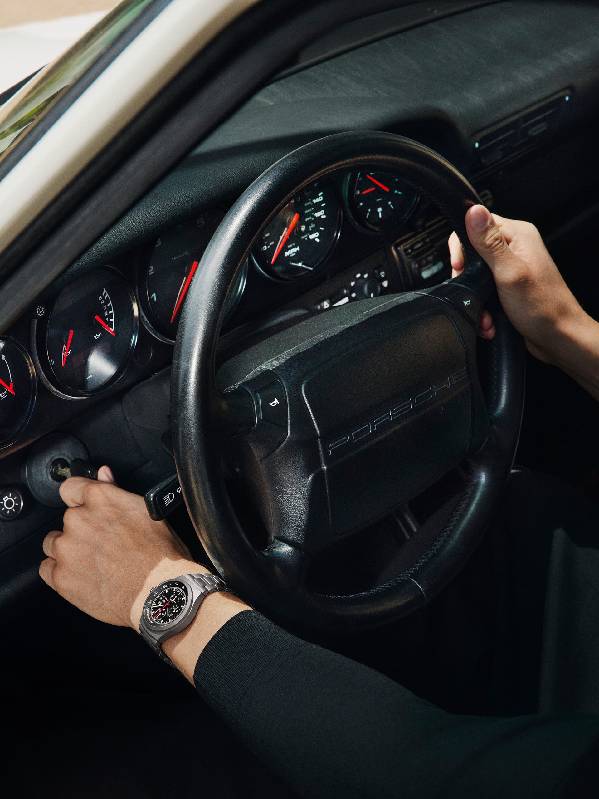 Hands on Porsche steering wheel, dashboard gauges visible, person wearing a silver watch, car interior in black