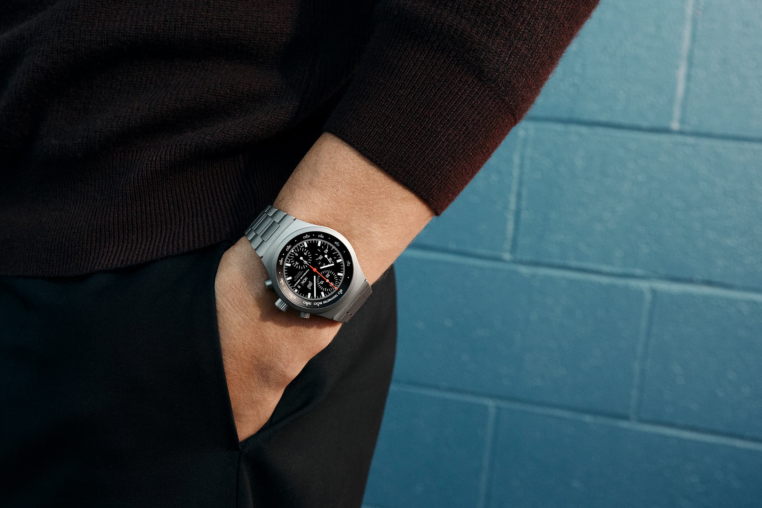 Silberne Chronographenarmbanduhr mit schwarzem Zifferblatt am Handgelenk, getragen mit dunklem Pullover vor blauen Fliesen