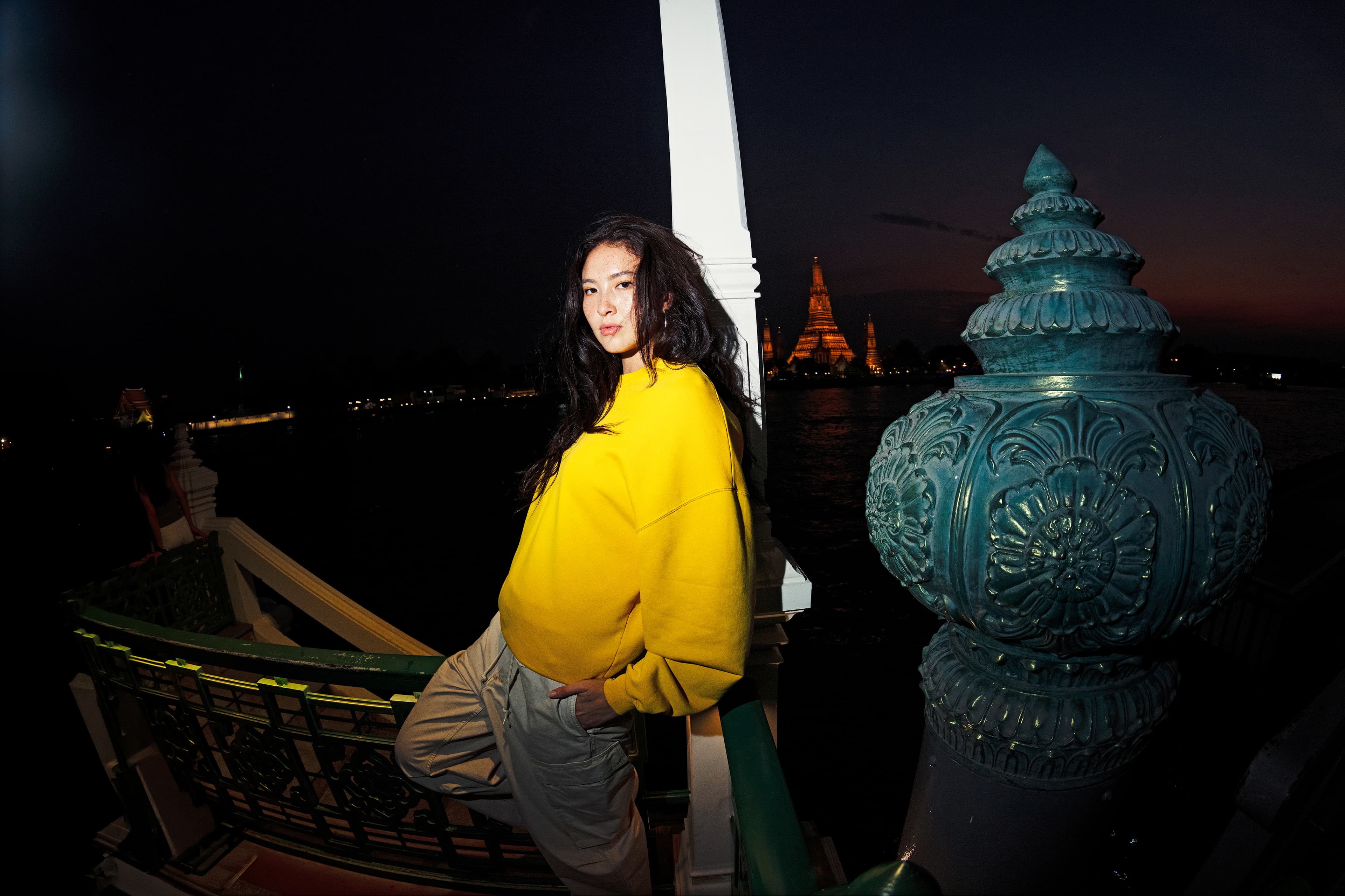 Mujer con sudadera Porsche AHEAD amarilla y pantalones beige en unas escaleras junto al río por la noche, con luces de un templo al fondo.