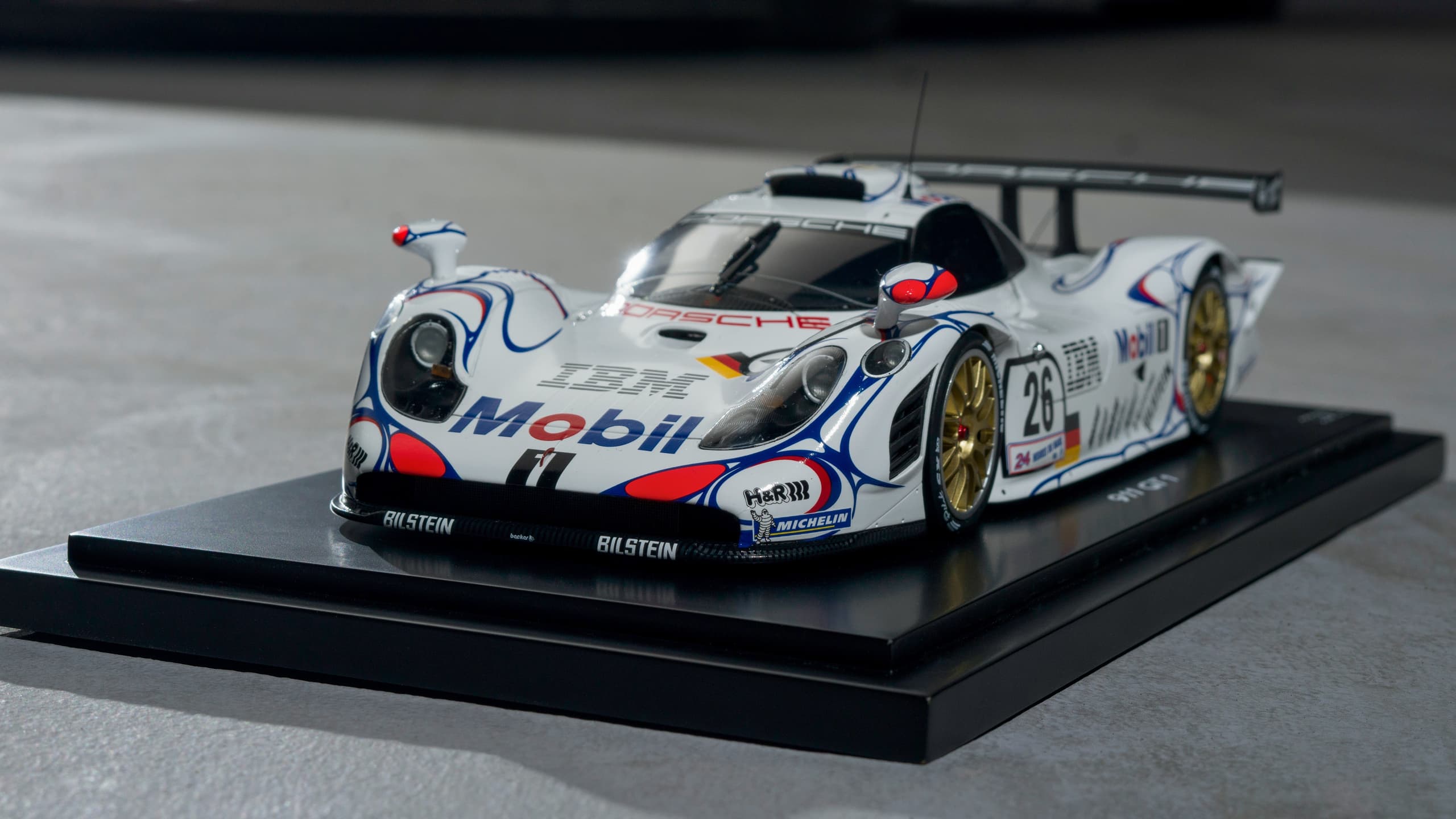 Modelo de carro de corrida Porsche branco com decalques coloridos e rodas douradas em suporte de exibição preto.