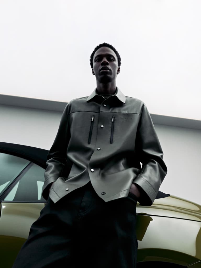 Man in gray jacket stands confidently beside a green car, under a cloudy sky.