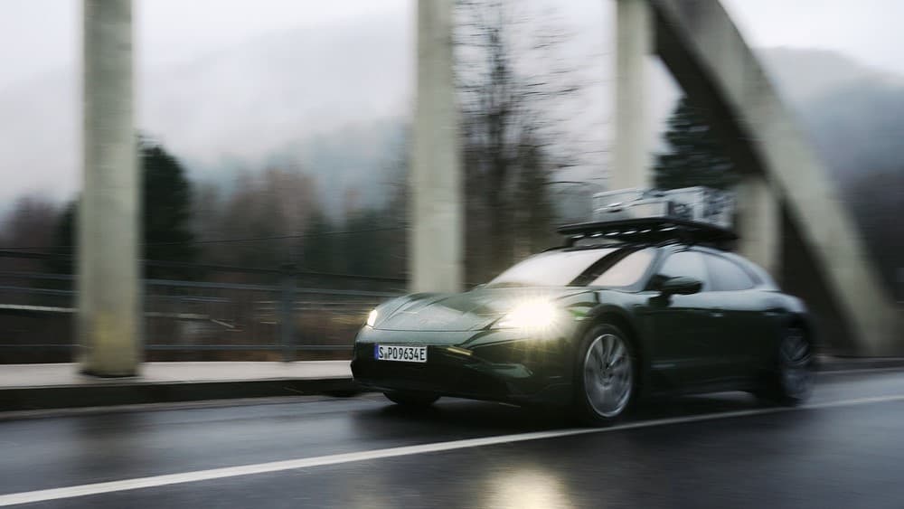 Grüner Porsche mit Dachgepäckträger fährt auf nasser Straße, Scheinwerfer an, unter einer Brücke in nebliger, bewaldeter Gegend.