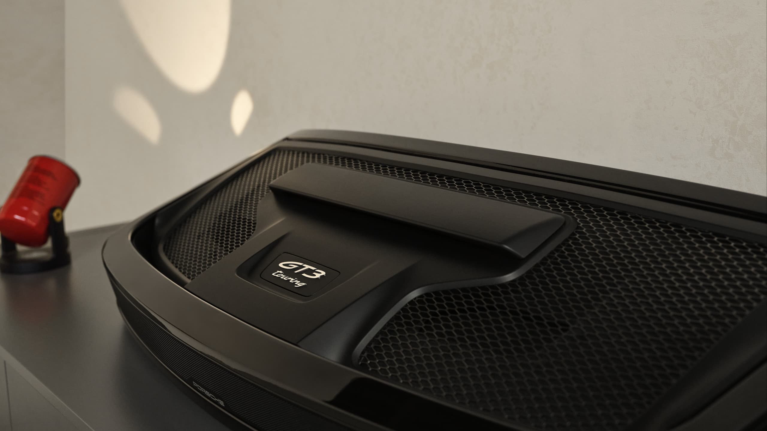 Black Porsche GT3 touring engine cover on display, with a red fire extinguisher in the background on a gray surface.