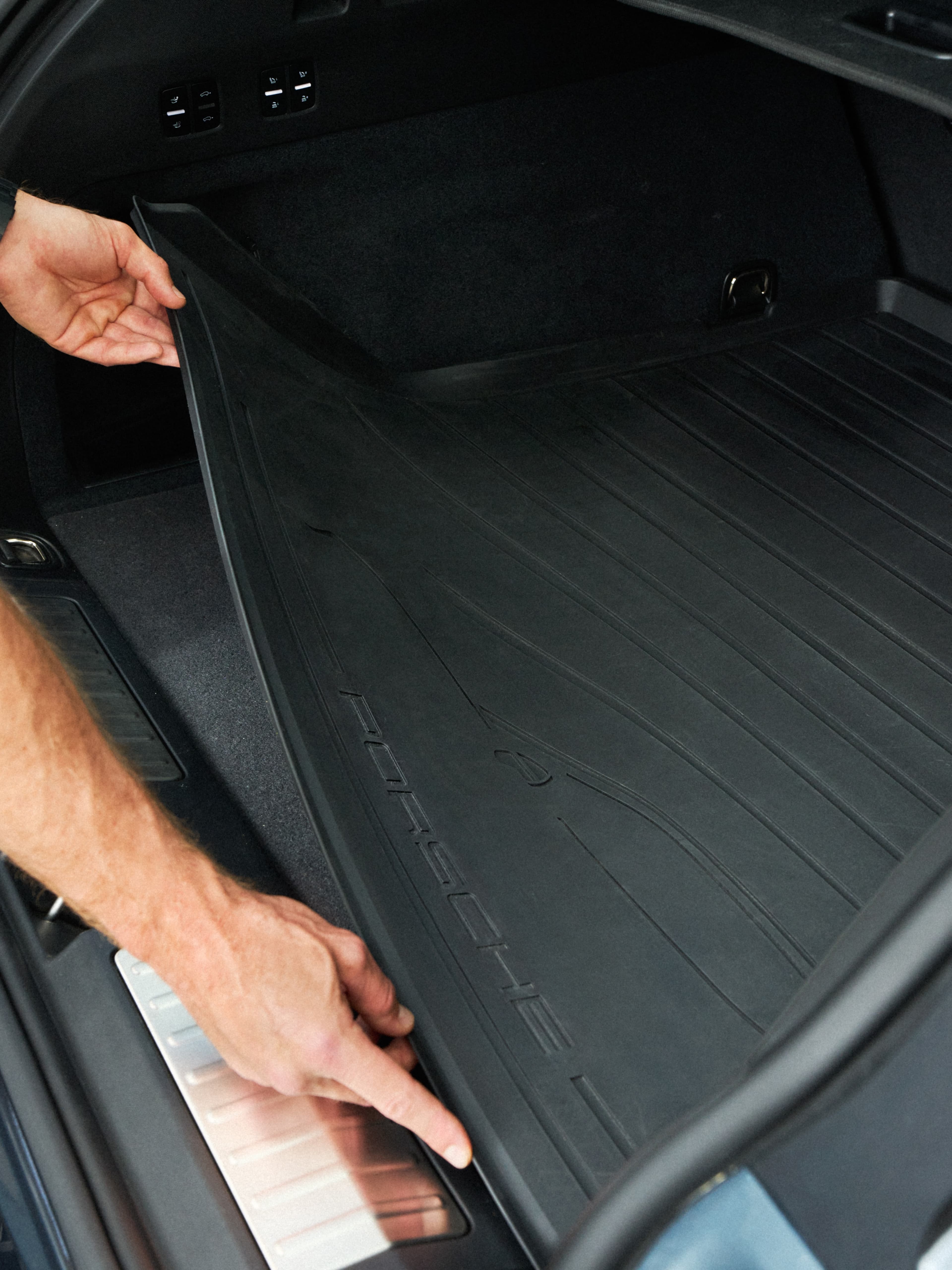 Hand placing black rubber trunk liner into a Porsche cargo area, close-up of textured mat and rear sill.