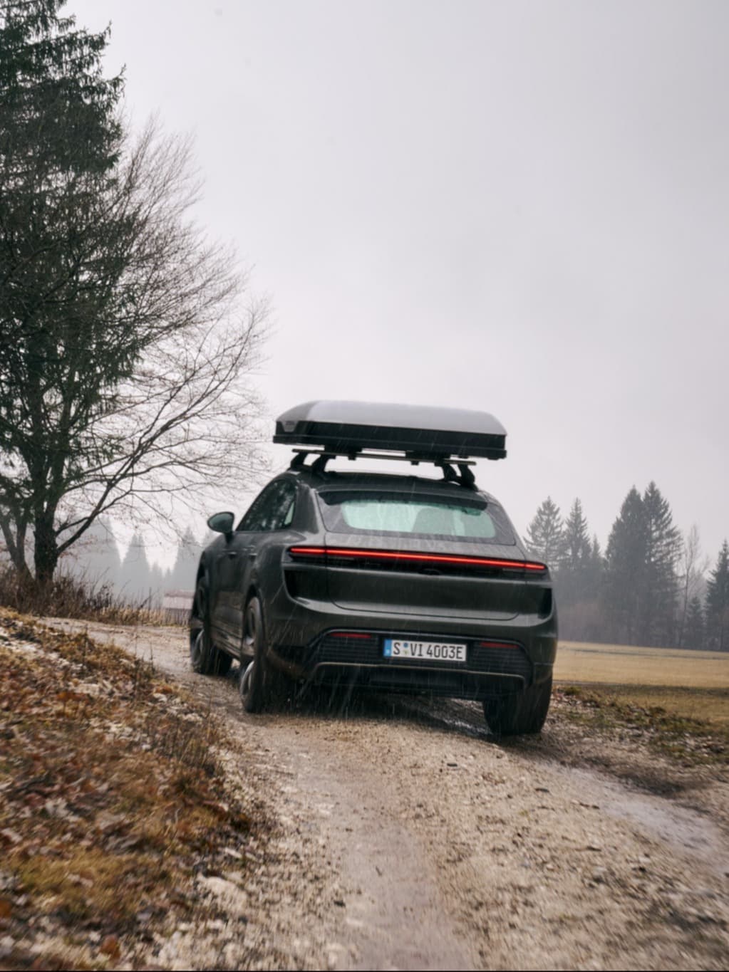 Porsche car with roof box driving on a muddy path, surrounded by trees on a cloudy day