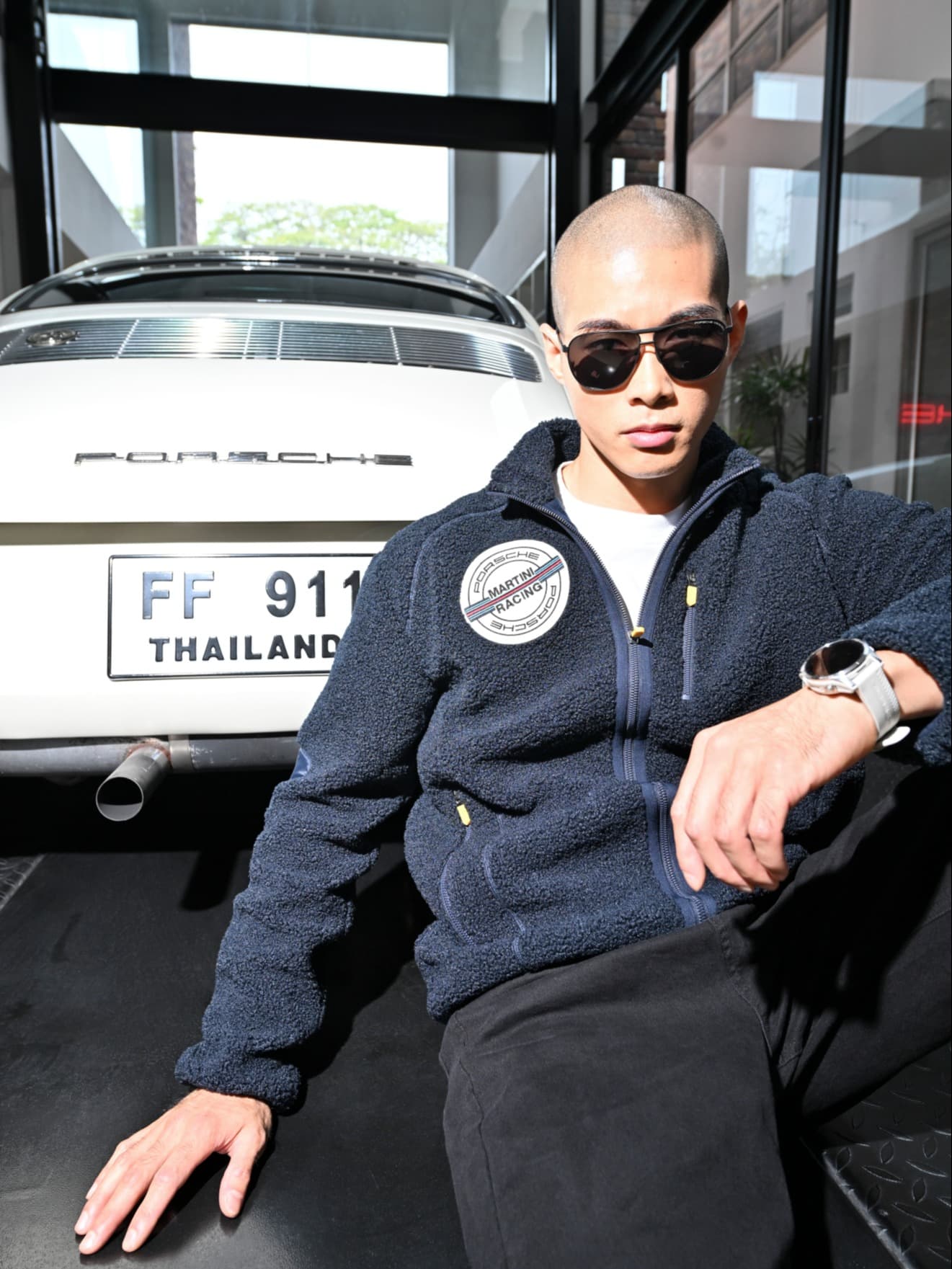 Man in sunglasses and navy jacket sits in front of a white Porsche with Thailand license plate, indoors.