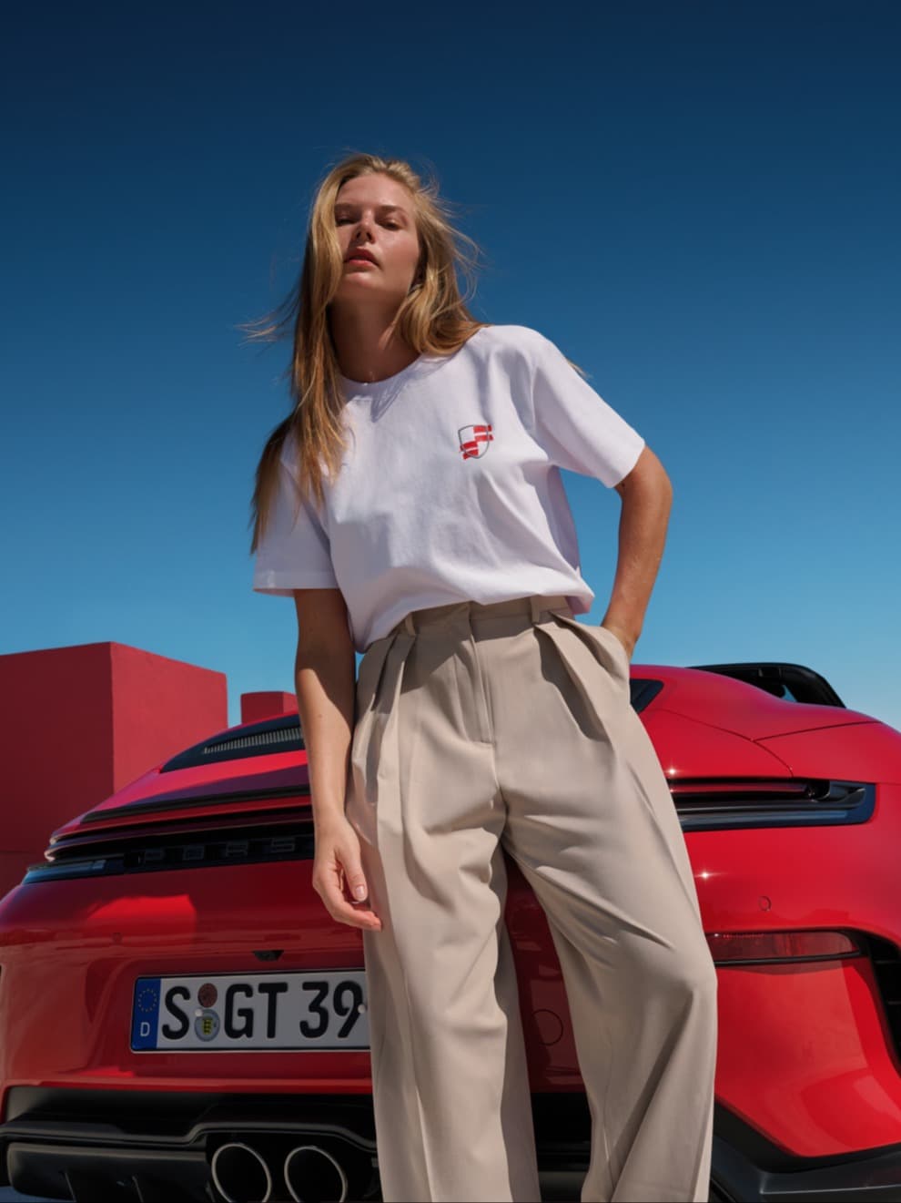 Vrouw in een wit T-shirt en beige broek, leunend tegen een rode Porsche cabriolet onder een blauwe lucht.