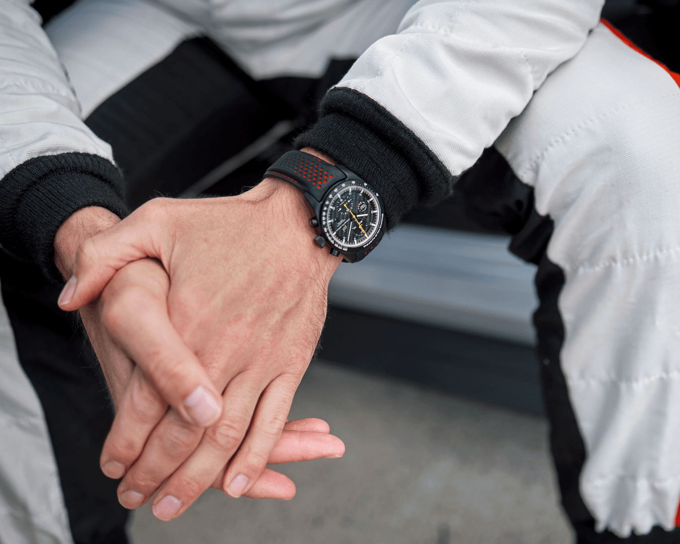 Na zdjęciu siedzący mężczyzna w kombinezonie wyścigowym, z dłońmi splecionymi, ma na nadgarstku zegarek Porsche Chronograph 1 GT3 RS.