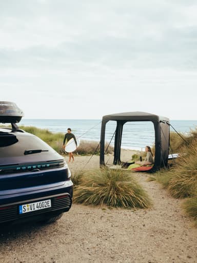 SUV Porsche avec coffre de toit, garé près des dunes ; un surfeur portant sa planche passe devant une tente de plage noire au bord de la mer.
