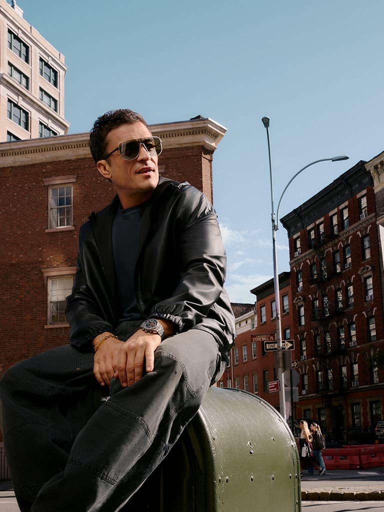 Homem de óculos de sol e casaco preto sentado numa caixa de correio verde em ambiente urbano, com edifícios de tijolo e céu limpo ao fundo.