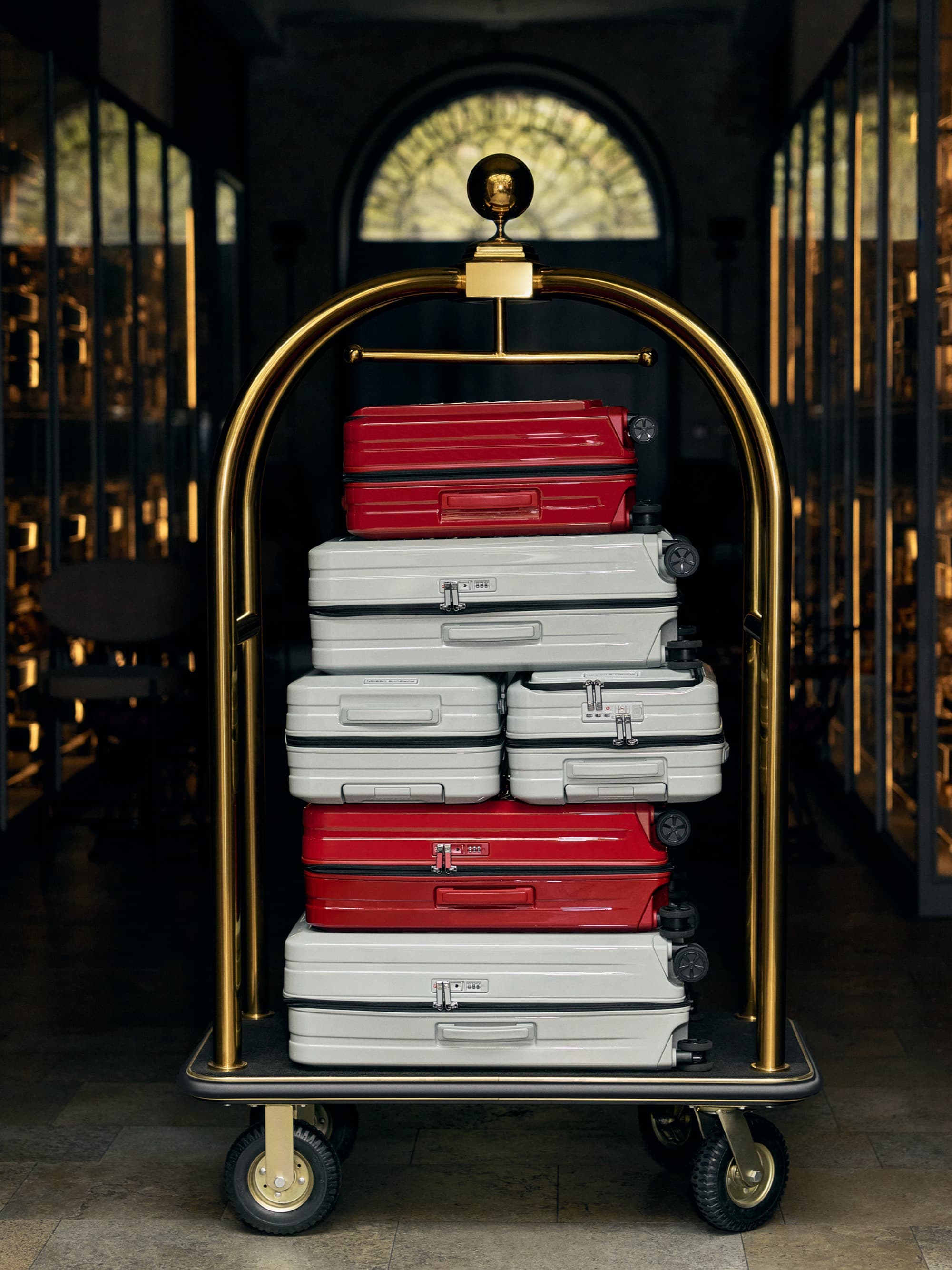 Gold luggage cart with stacked red and white suitcases in a dimly lit hallway with arched window in the background.
