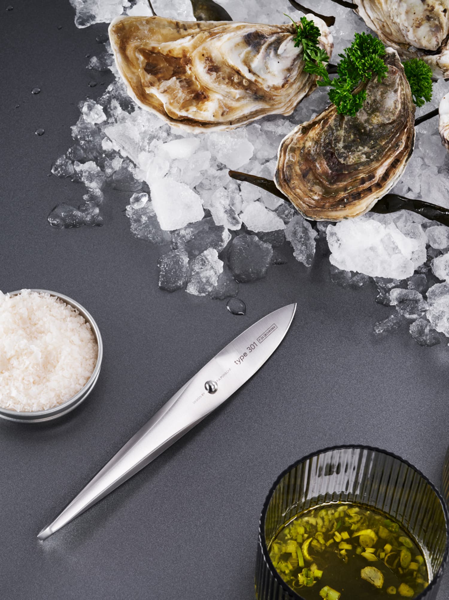 Oysters on ice with parsley, a sleek silver knife, a bowl of salt, and a glass of oil with herbs on a dark surface.