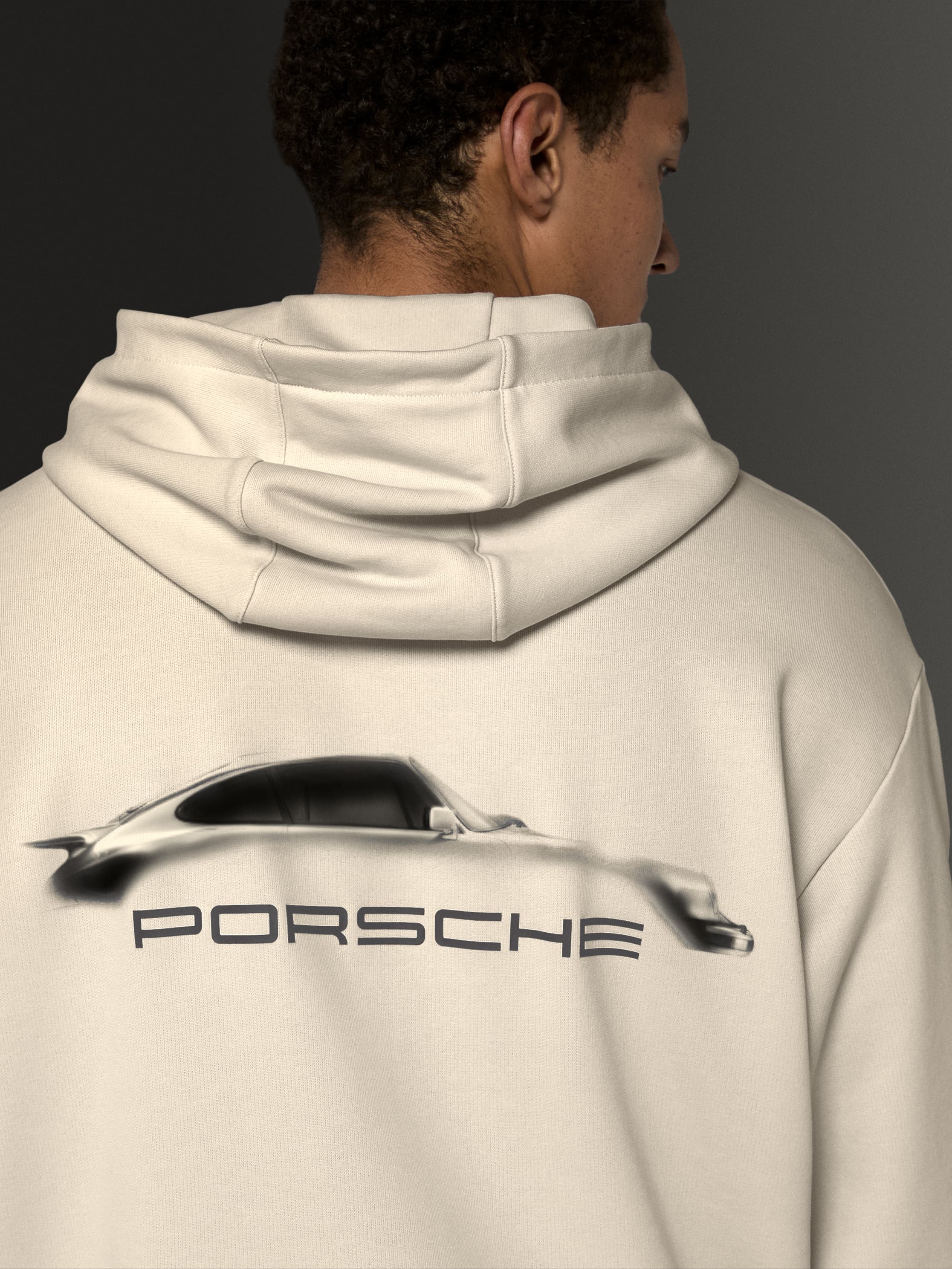 Man wearing cream hoodie with Porsche car silhouette and logo on back, against a dark background