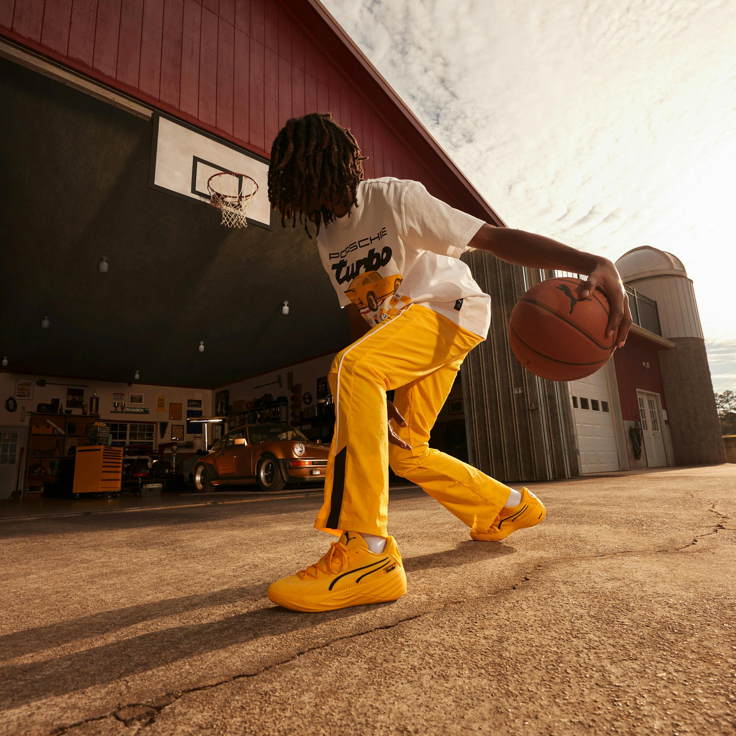 The image depicts a man playing basketball outdoors. He is wearing yellow Porsche x Puma clothing and appears to be on a basketball court. The image also includes a Porsche Turbo car