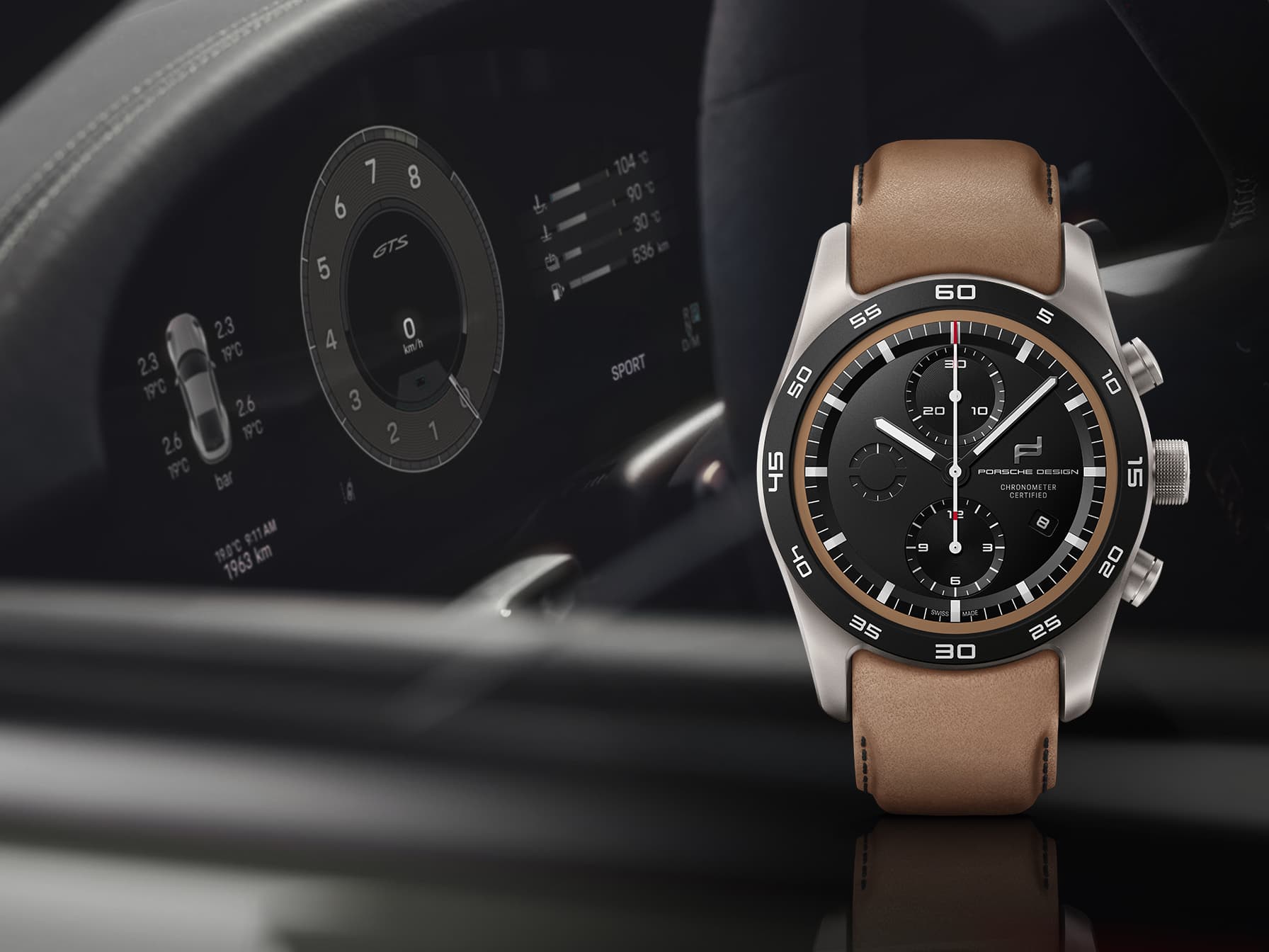 Porsche Design Chronograph watch with black dial and brown leather strap, in front of a blurred Porsche dashboard.