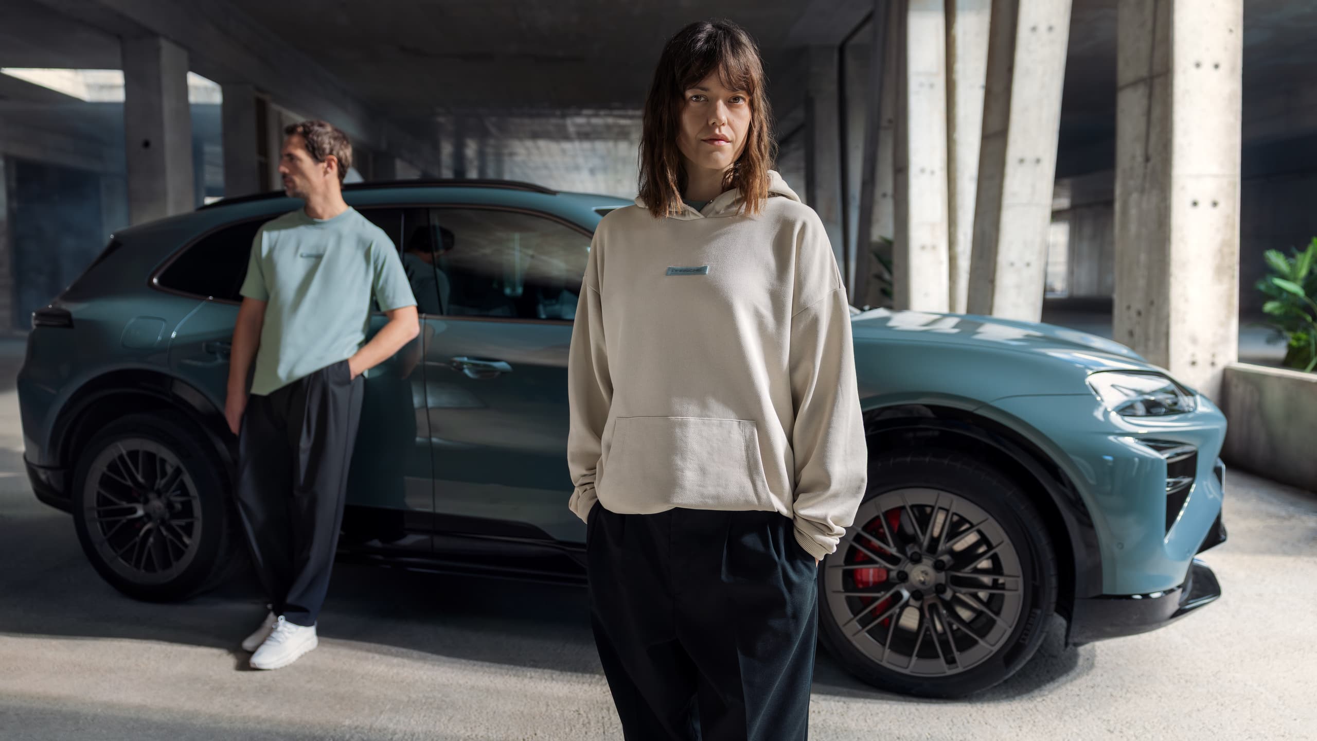 Two people stand near a blue Porsche SUV in a concrete garage, wearing casual clothing.