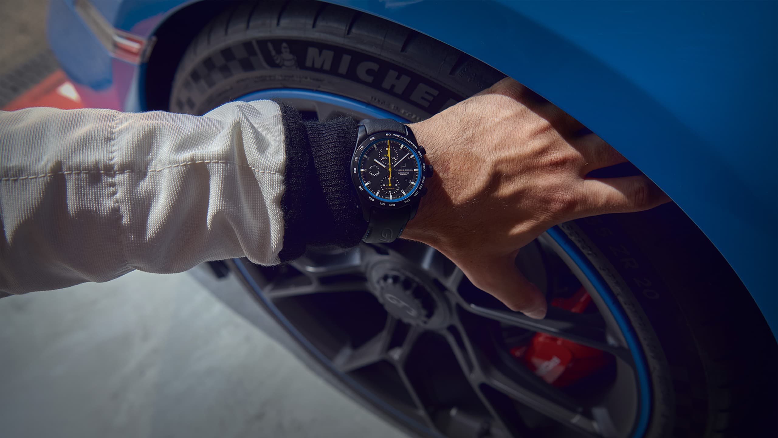 Na zdjęciu zegarek na rękę Porsche Chronograph 911 GT3, noszony przez mężczyznę sięgającego z góry do opony samochodu Porsche.