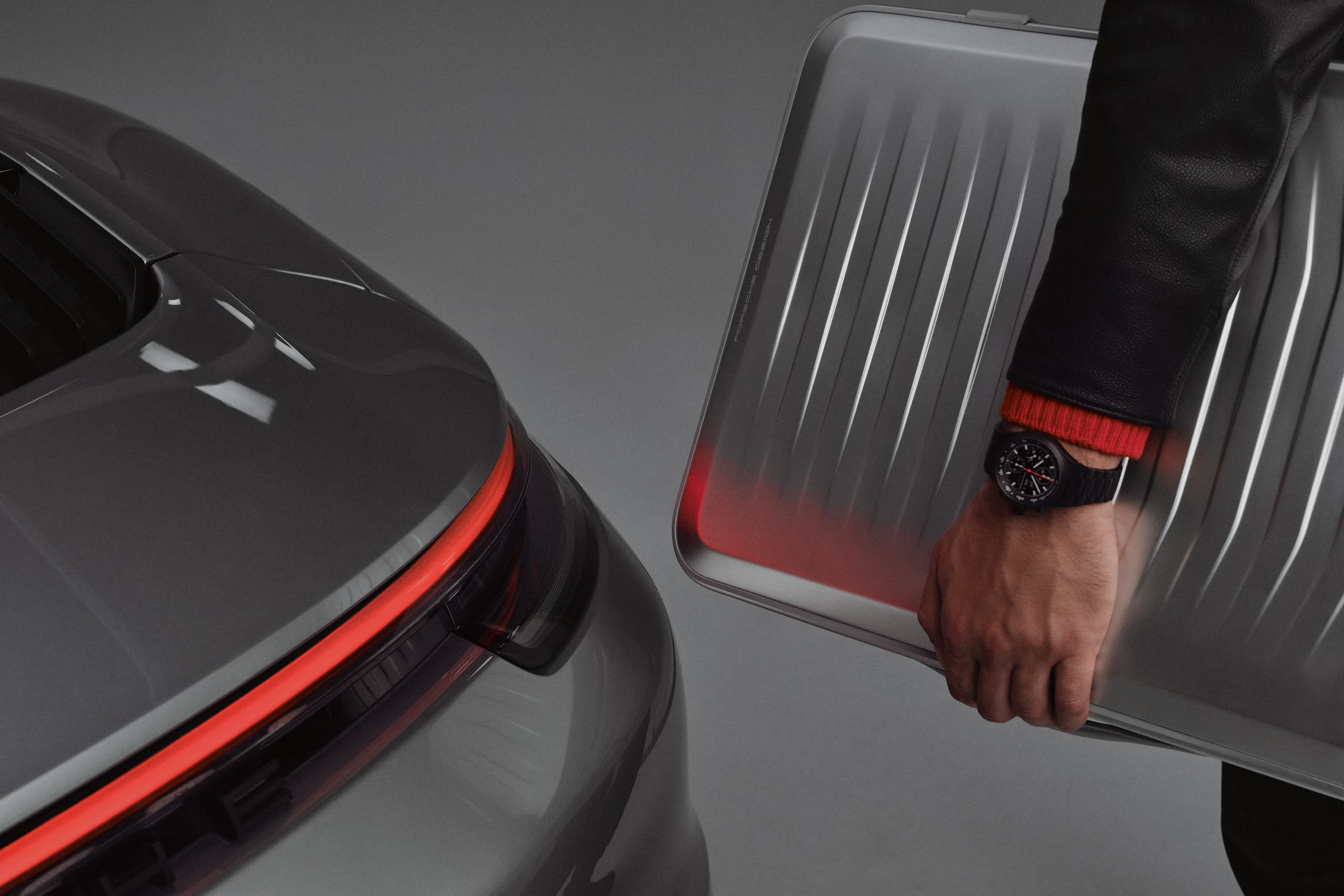 Dłoń trzyma srebrną walizkę Porsche Design obok szarego tyłu Porsche z czerwonym pasem tylnych świateł.