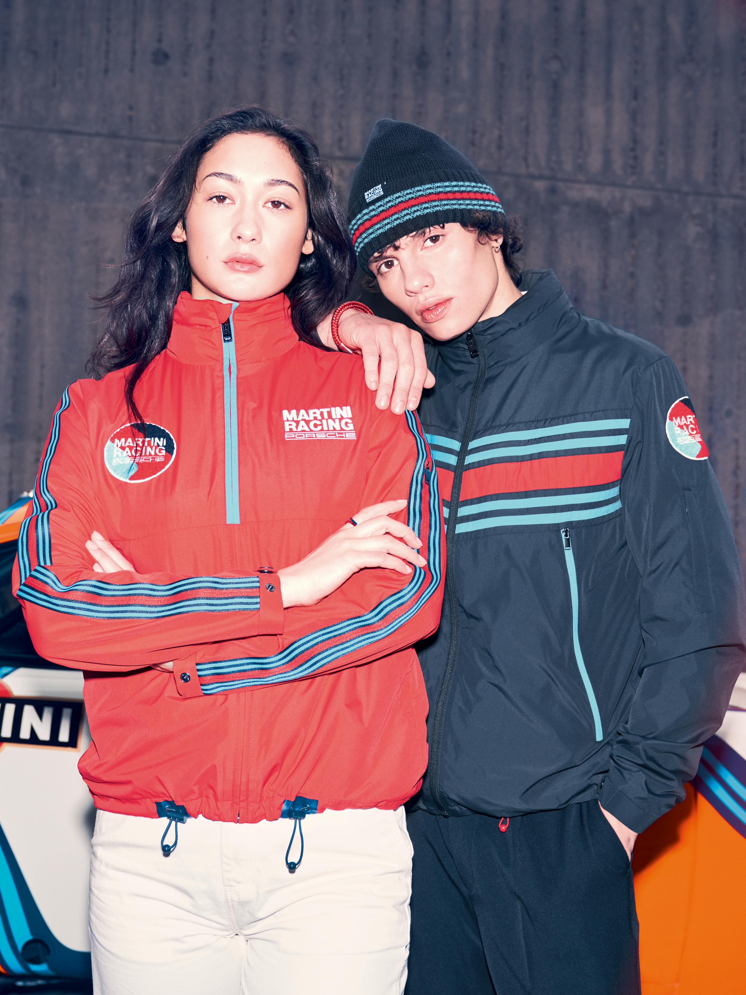 A woman and a man stand next to each other wearing Porsche MARTINI RACING jackets and a basket