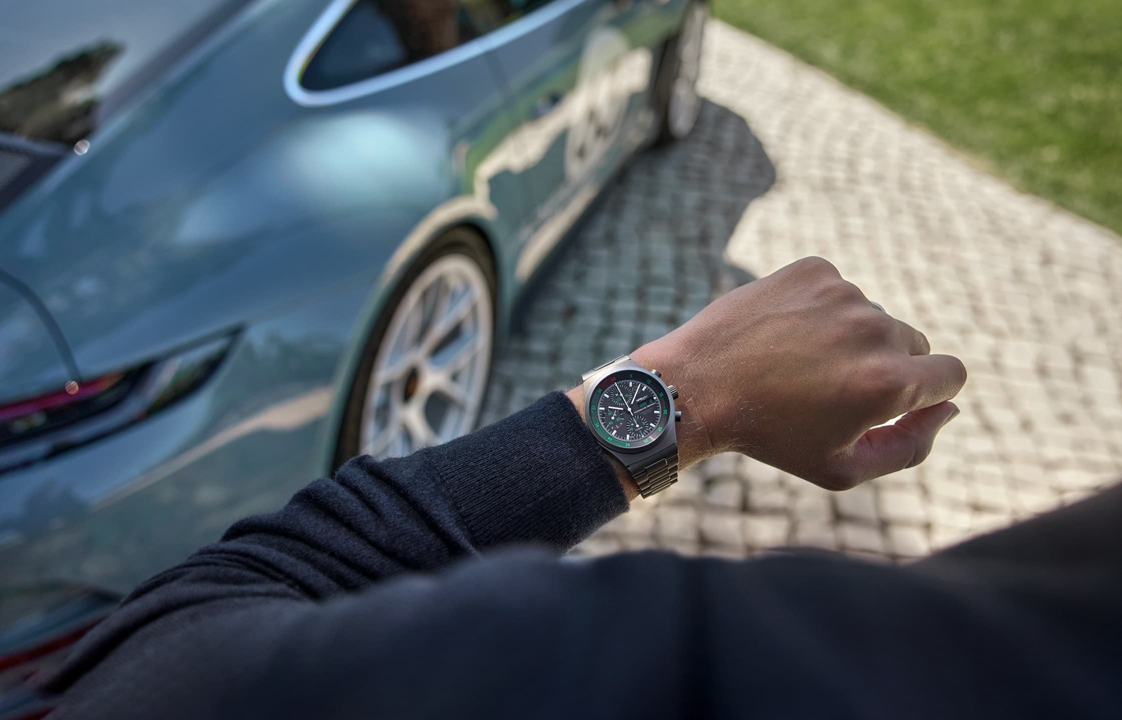 Silver wristwatch from Porsche on the wrist of a man, photographed from above. A light blue Porsche can be seen cropped in the background