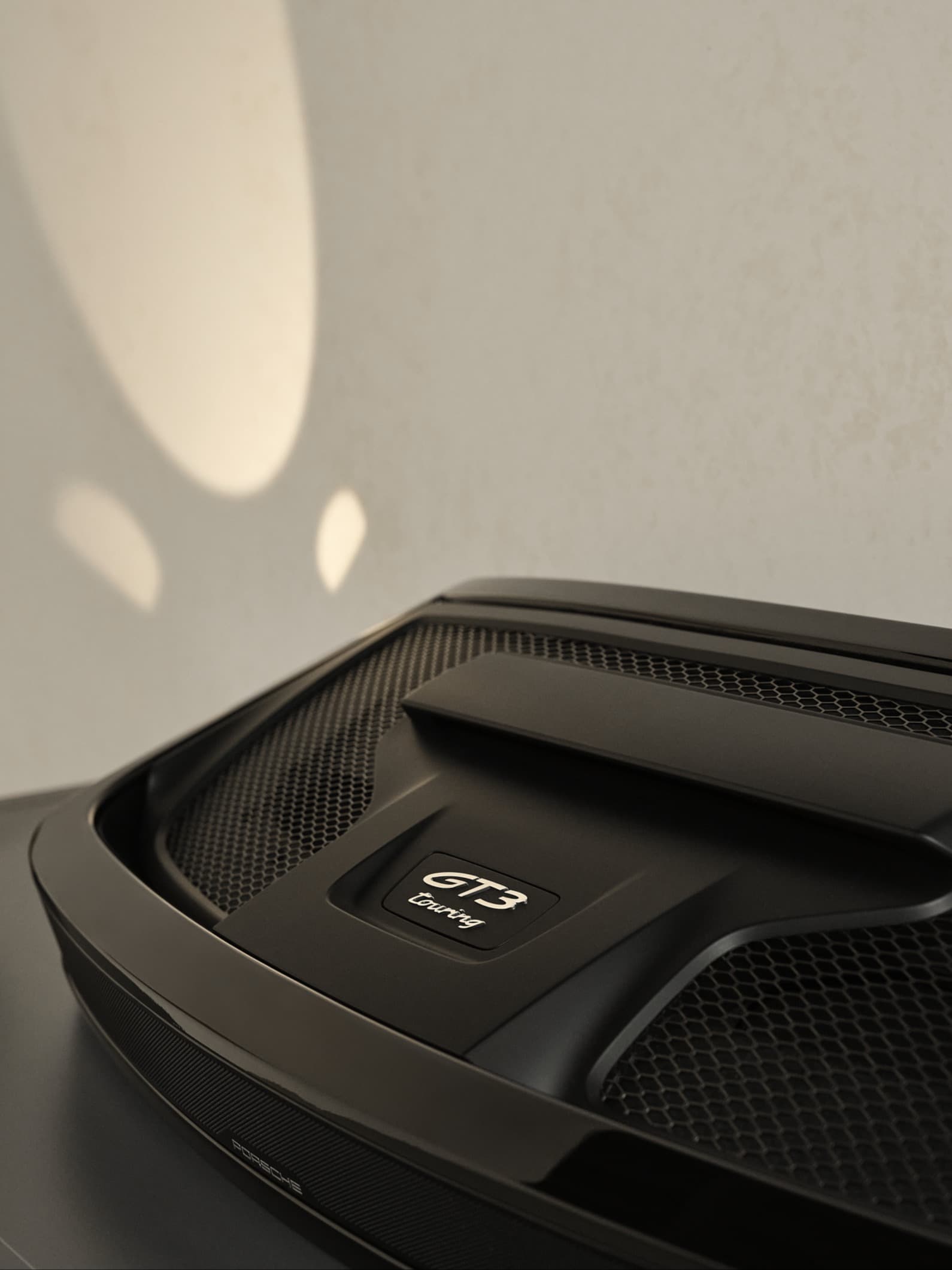 Nahaufnahme des schwarzen Porsche GT3 Touring-Motorabdeckungs mit Mesh-Details, vor einer hellen Wand mit weichen Schatten.