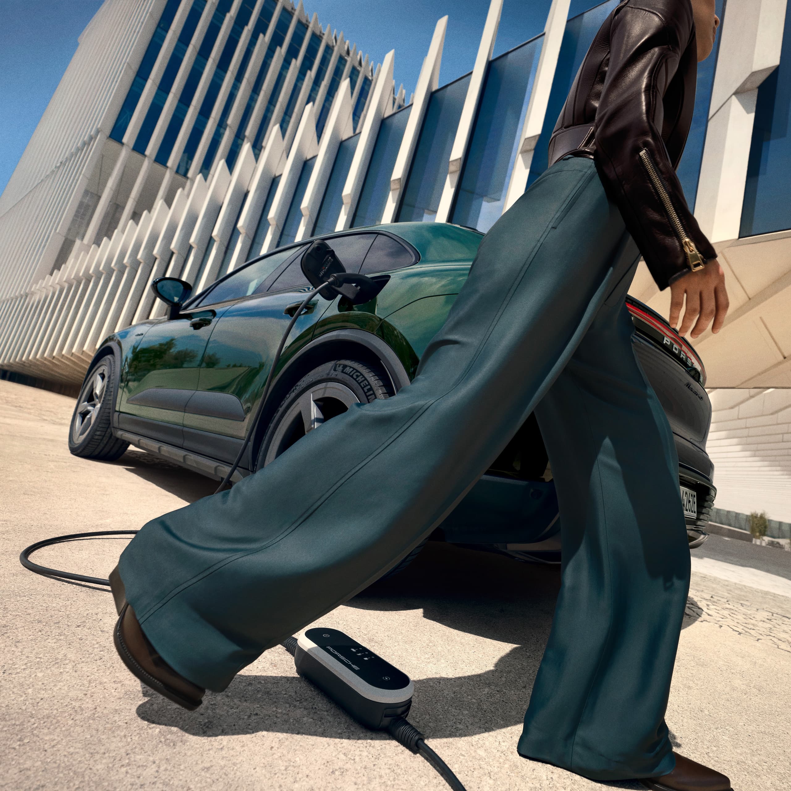 Woman is walking in front of a darkgreen Porsche Macan with a mobile charger on the ground, modern building in the background