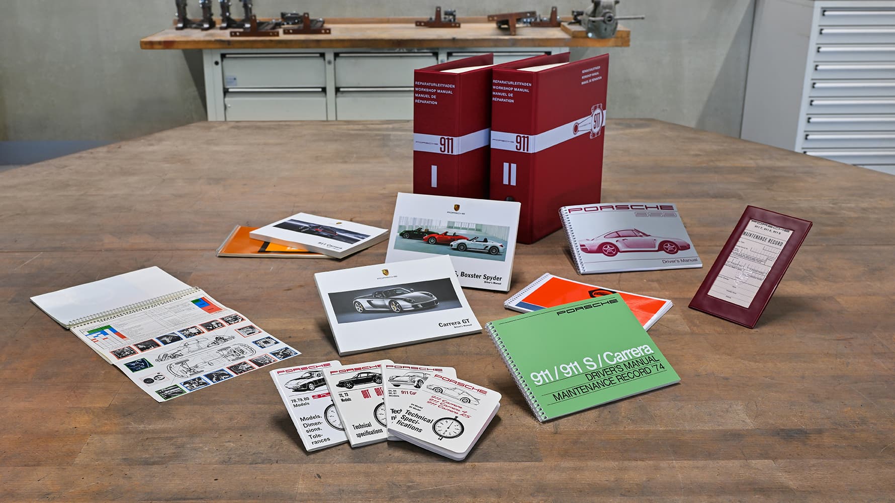 Porsche-Handbücher und Broschüren auf einem Holztisch, mit Modellen wie Carrera GT und Boxster Spyder, in roten Ordnern.