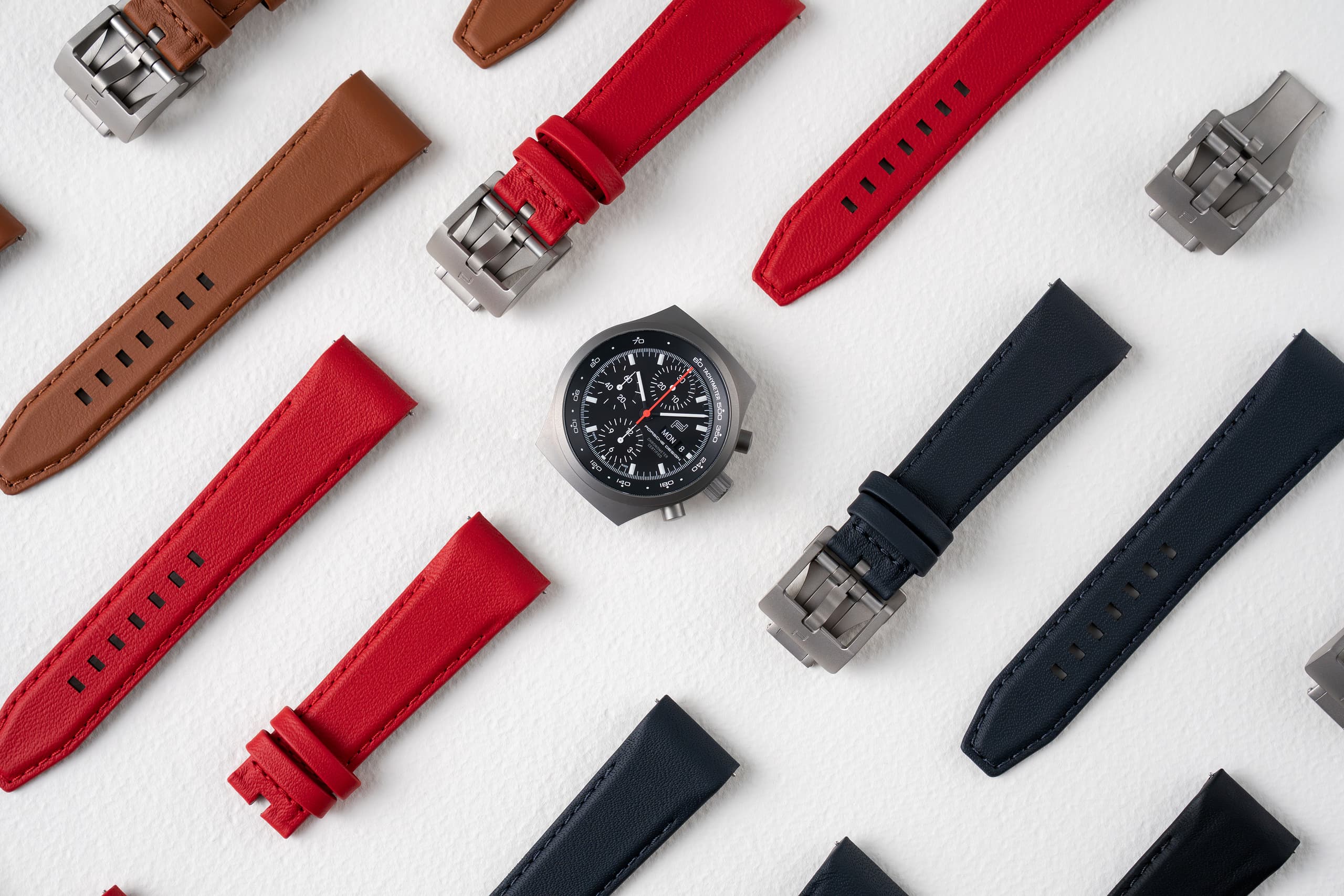 Chronograafhorloge met zwarte wijzerplaat, omringd door verwisselbare leren horlogebandjes in rood, bruin en marineblauw op een witte achtergrond.