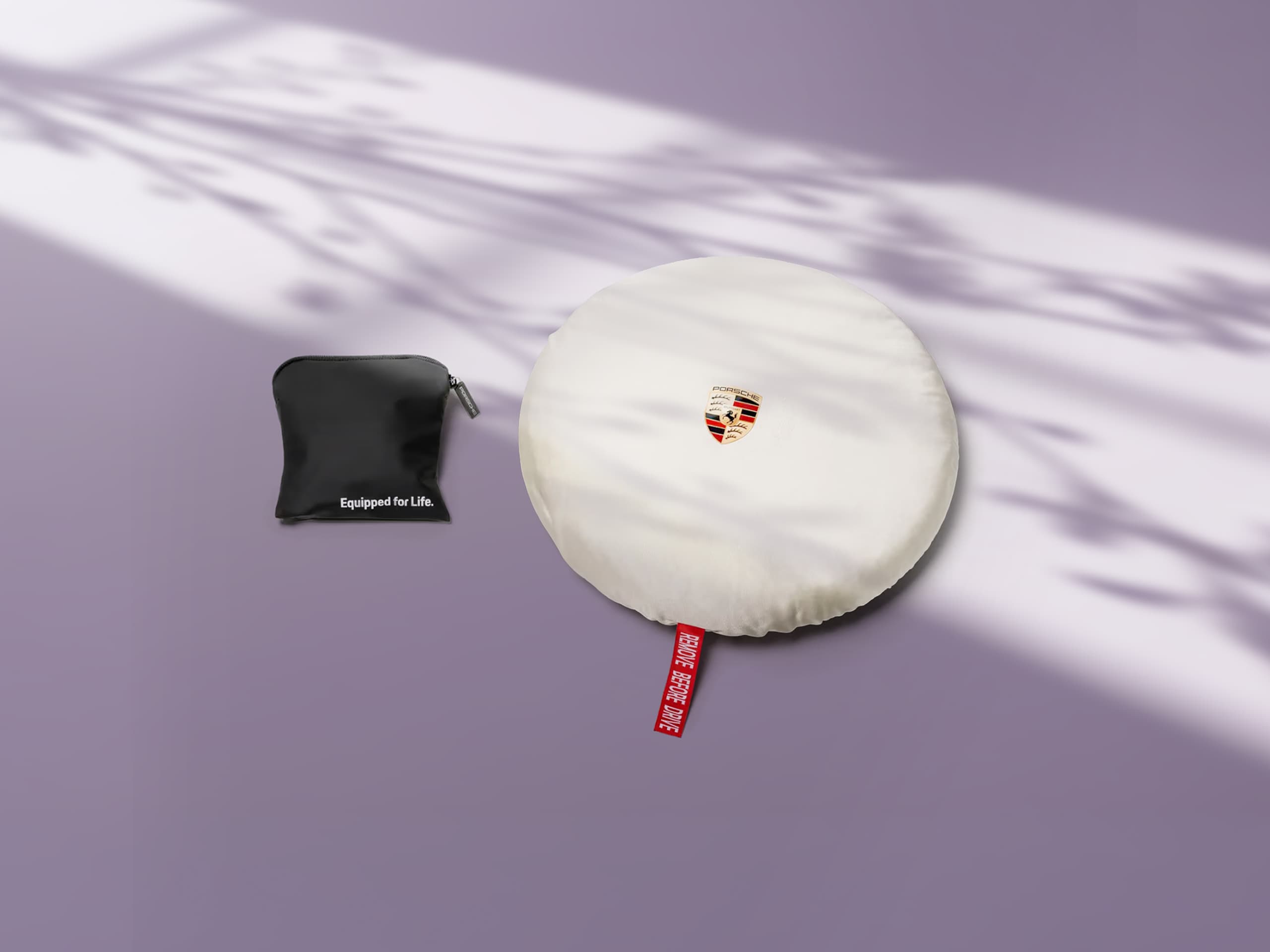 Hvidt Porsche-dæksel med logo og rød mærkat, ved siden af en sort pose på lilla baggrund med skyggeeffekter.