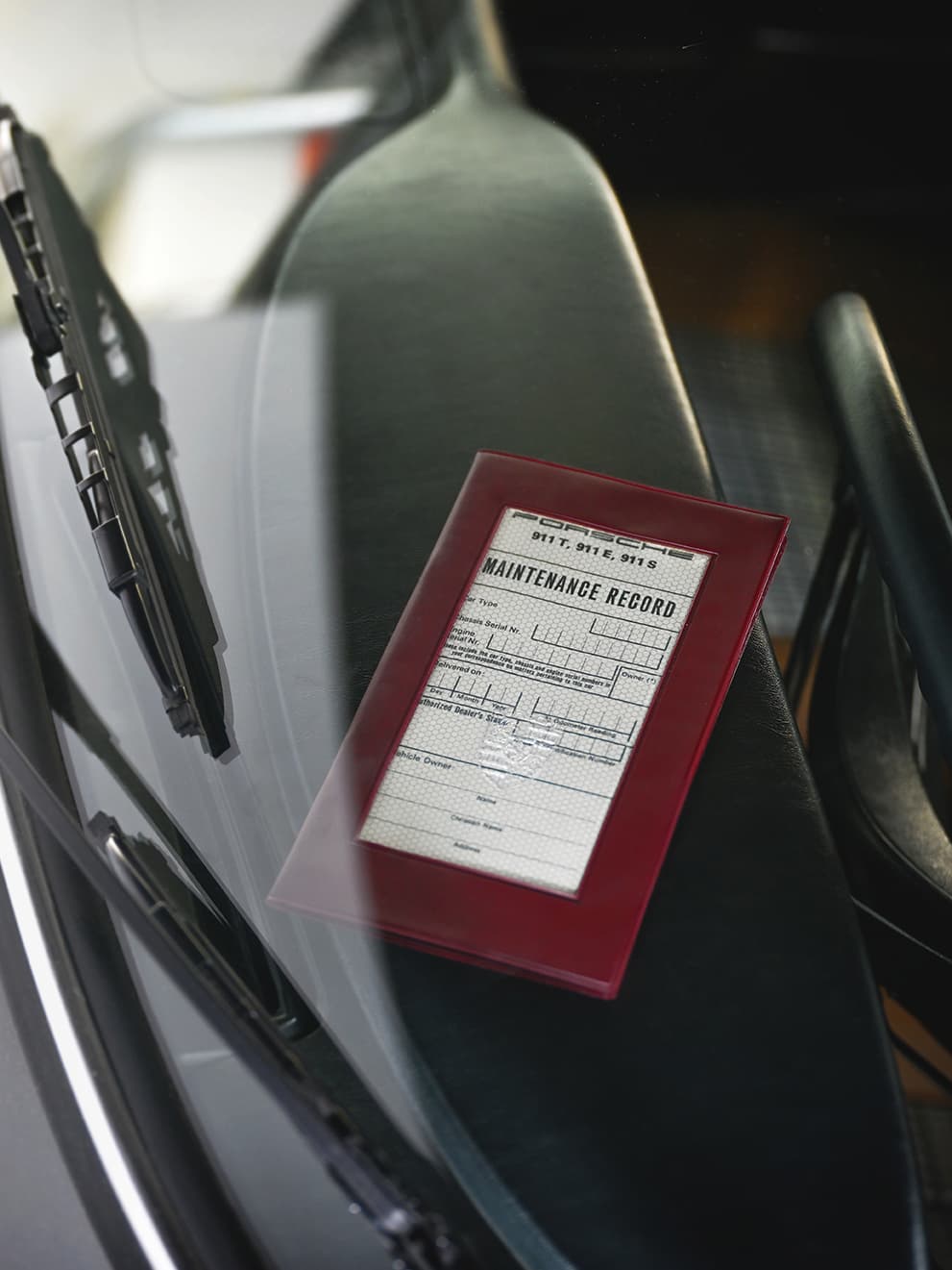 Maintenance record in red folder on car seat, viewed through windshield with wipers visible.