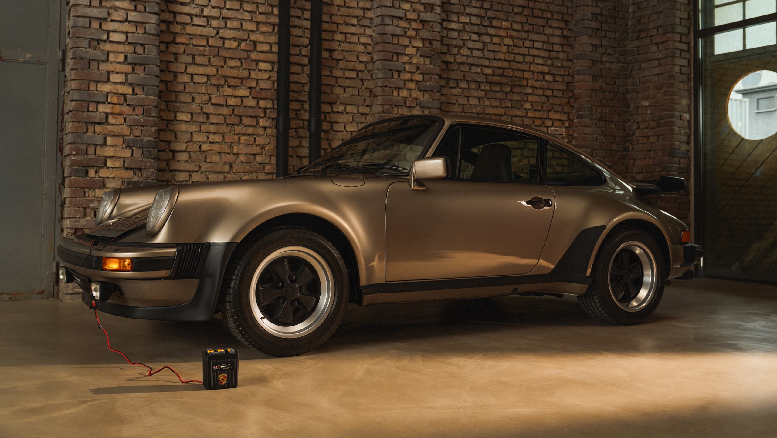 Klassischer goldener Porsche 911 in einer Backsteingarage, verbunden mit einem Batterieladegerät auf dem Boden.