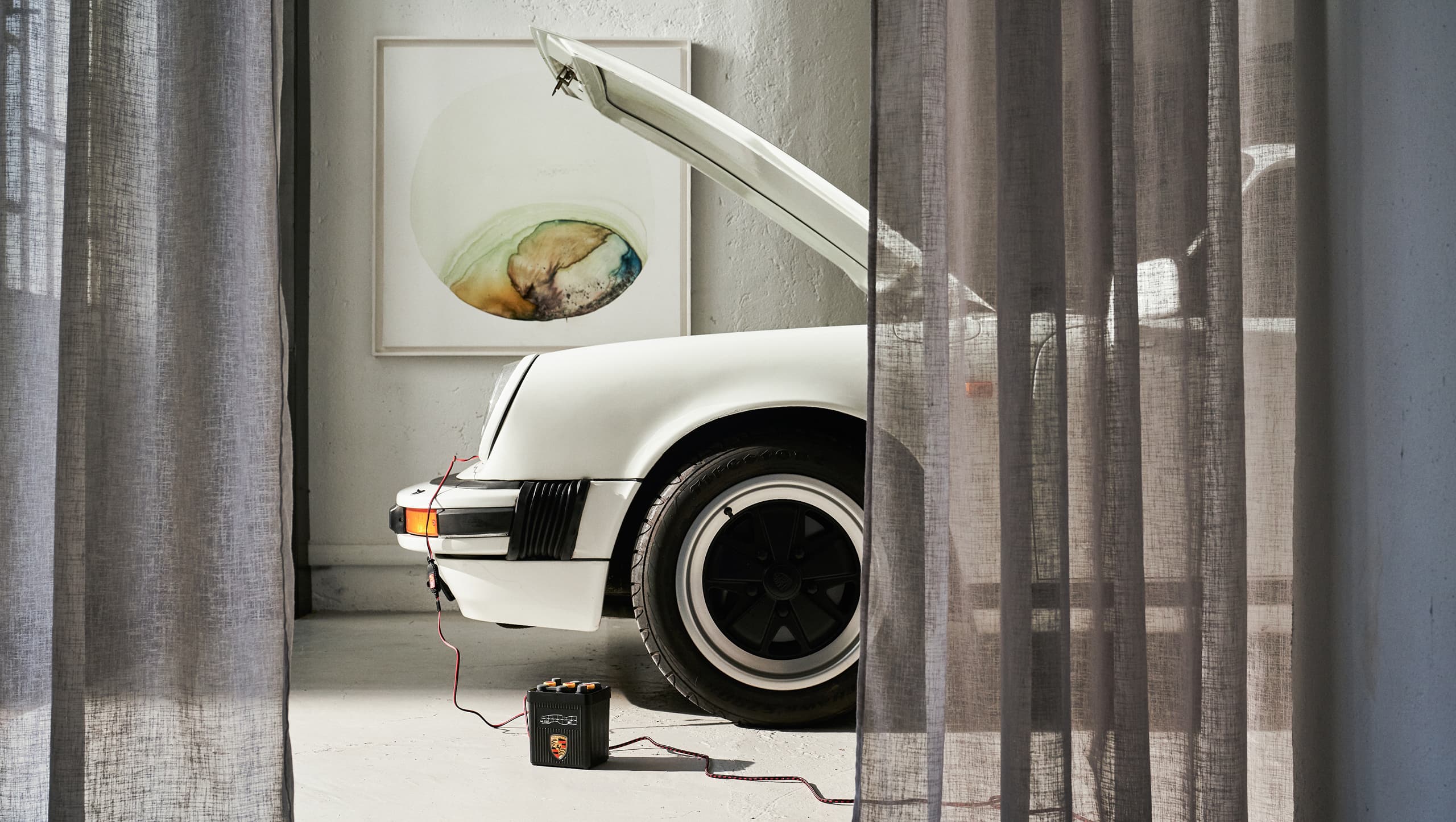 Weißer Porsche mit geöffnetem Motorhaube, angeschlossenem Batterieladegerät, hinter transparenten Vorhängen in einem hellen Raum mit abstrakter Wandkunst