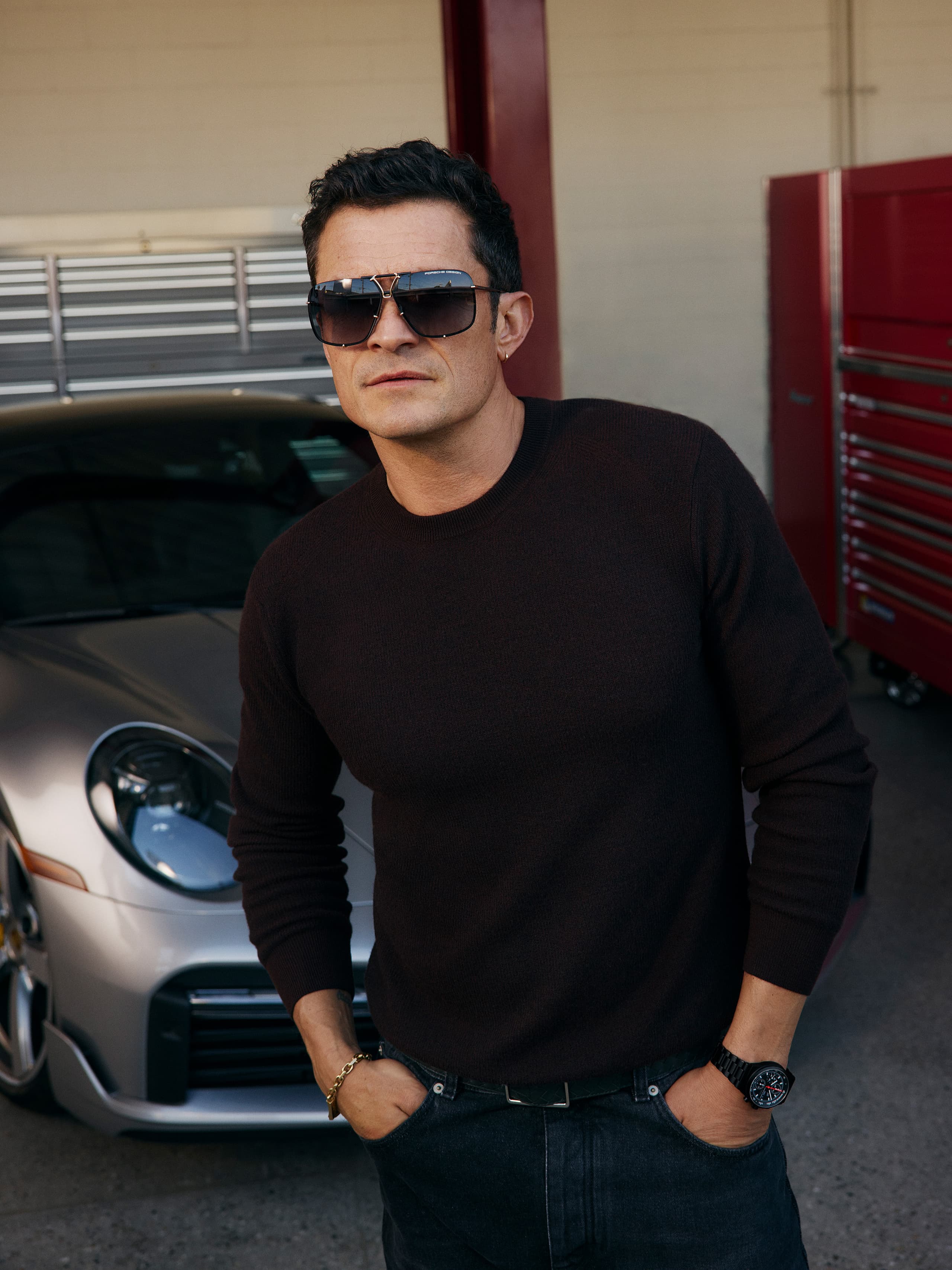 Orlando Bloom stands in a garage with a silver Porsche and wears sunglasses from Porsche Design.