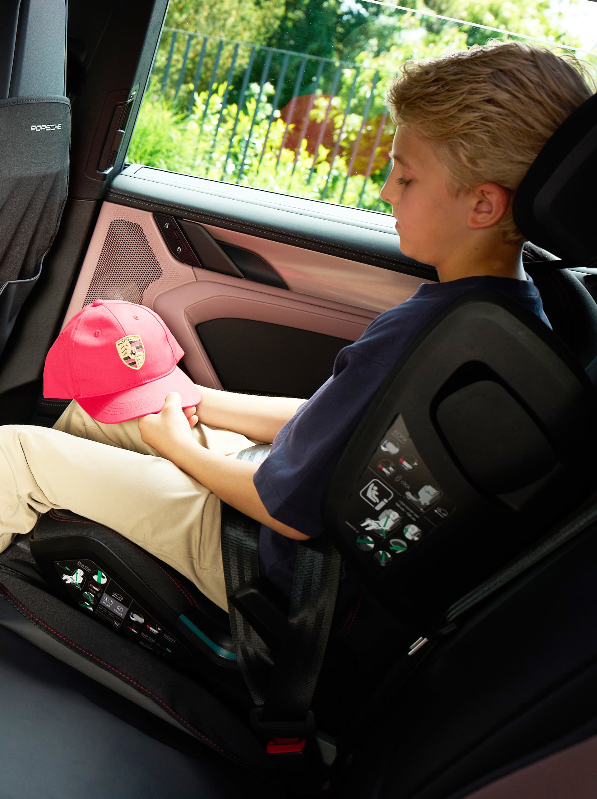Boy in a child car seat in the back seat, holding a red Porsche cap; view through the car window onto green scenery.