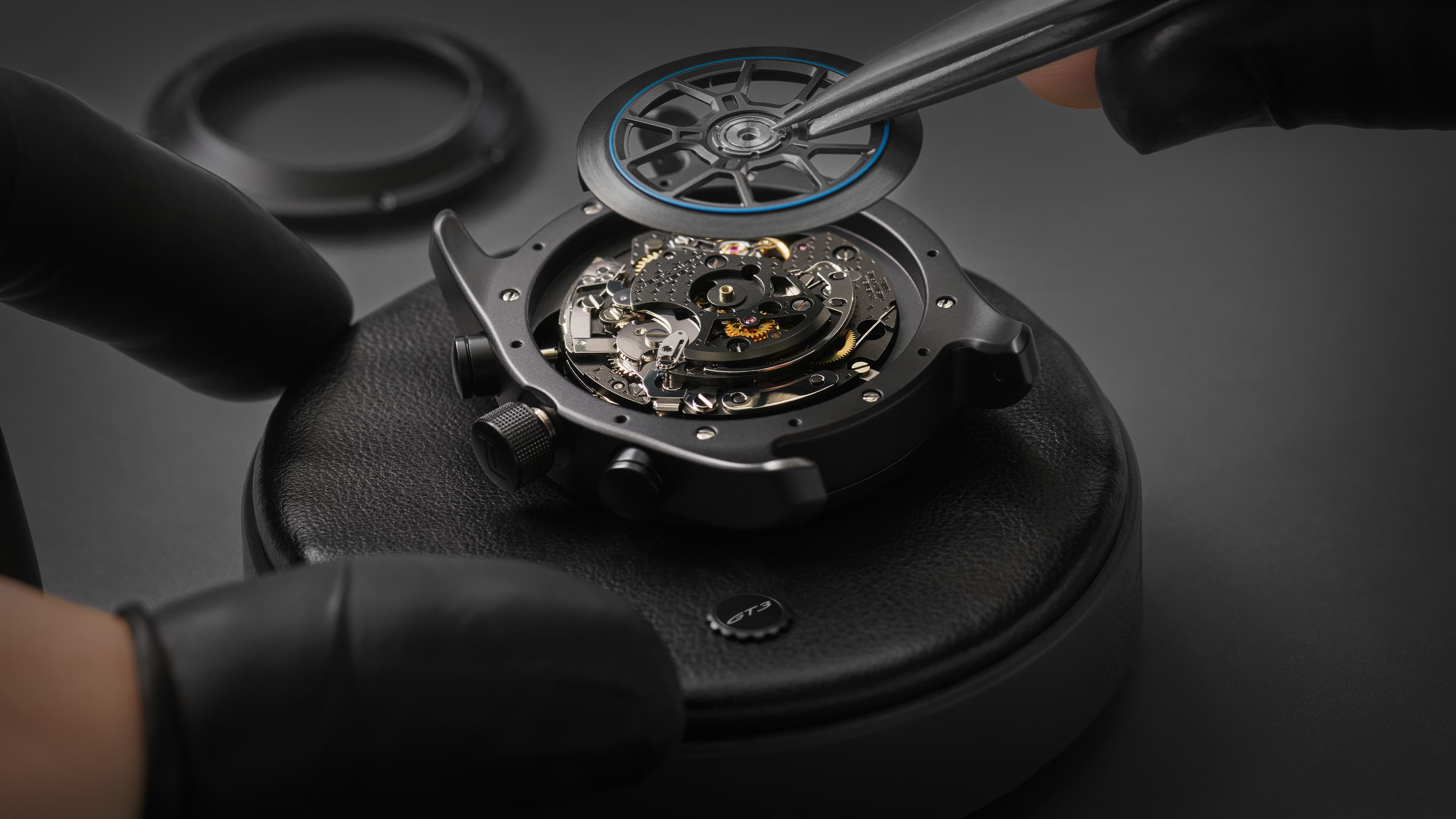 The movement in a black case, featuring a rotor with a rim design and a blue ring, is inserted using tweezers.